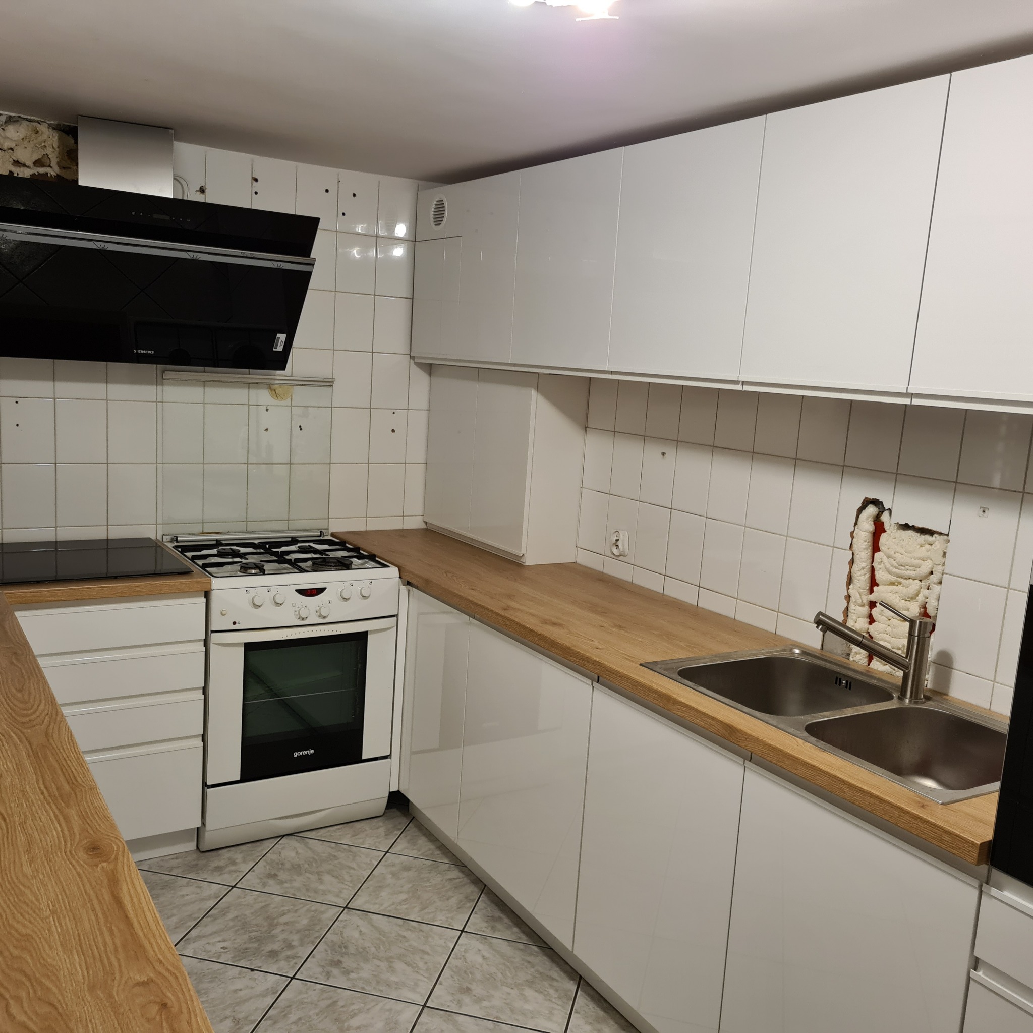 Biała kuchnia z drewnianym blatem, widoczna kuchenka gazowa, okap, zlew dwukomorowy i niedokończone wykończenie ściany za zlewem.