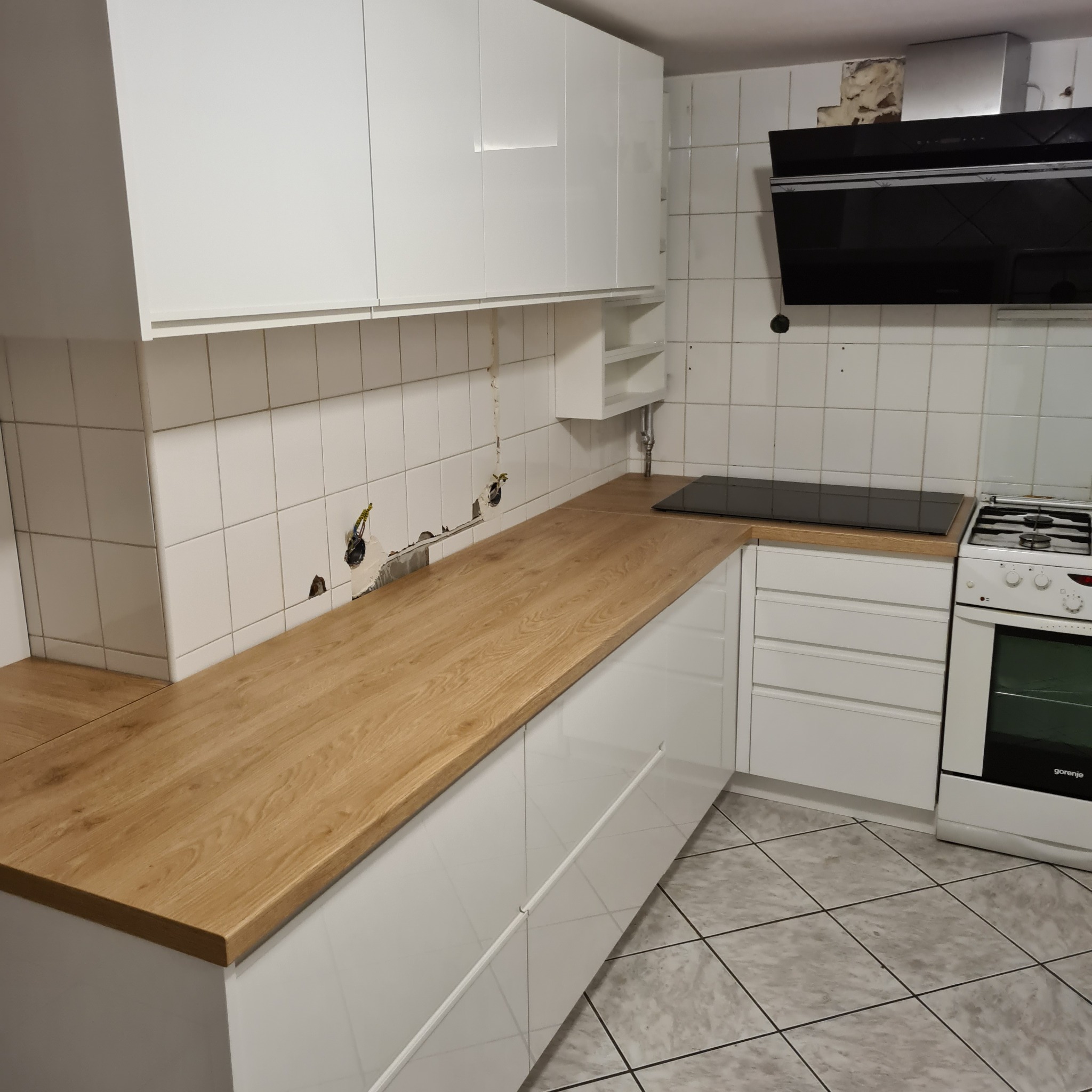 Montaż białych szafek kuchennych z drewnianym blatem, widoczne niedokończone prace przy instalacji elektrycznej i otwory w ścianie.