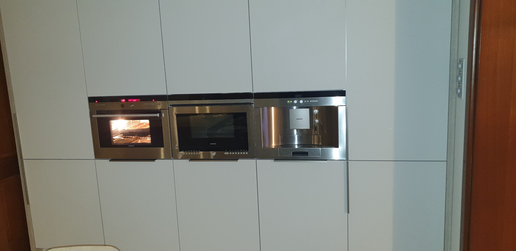 Nowoczesna zabudowa kuchenna z wbudowanym piekarnikiem, mikrofalówką i ekspresem do kawy marki Siemens, fronty białe, matowe, bez uchwytów.