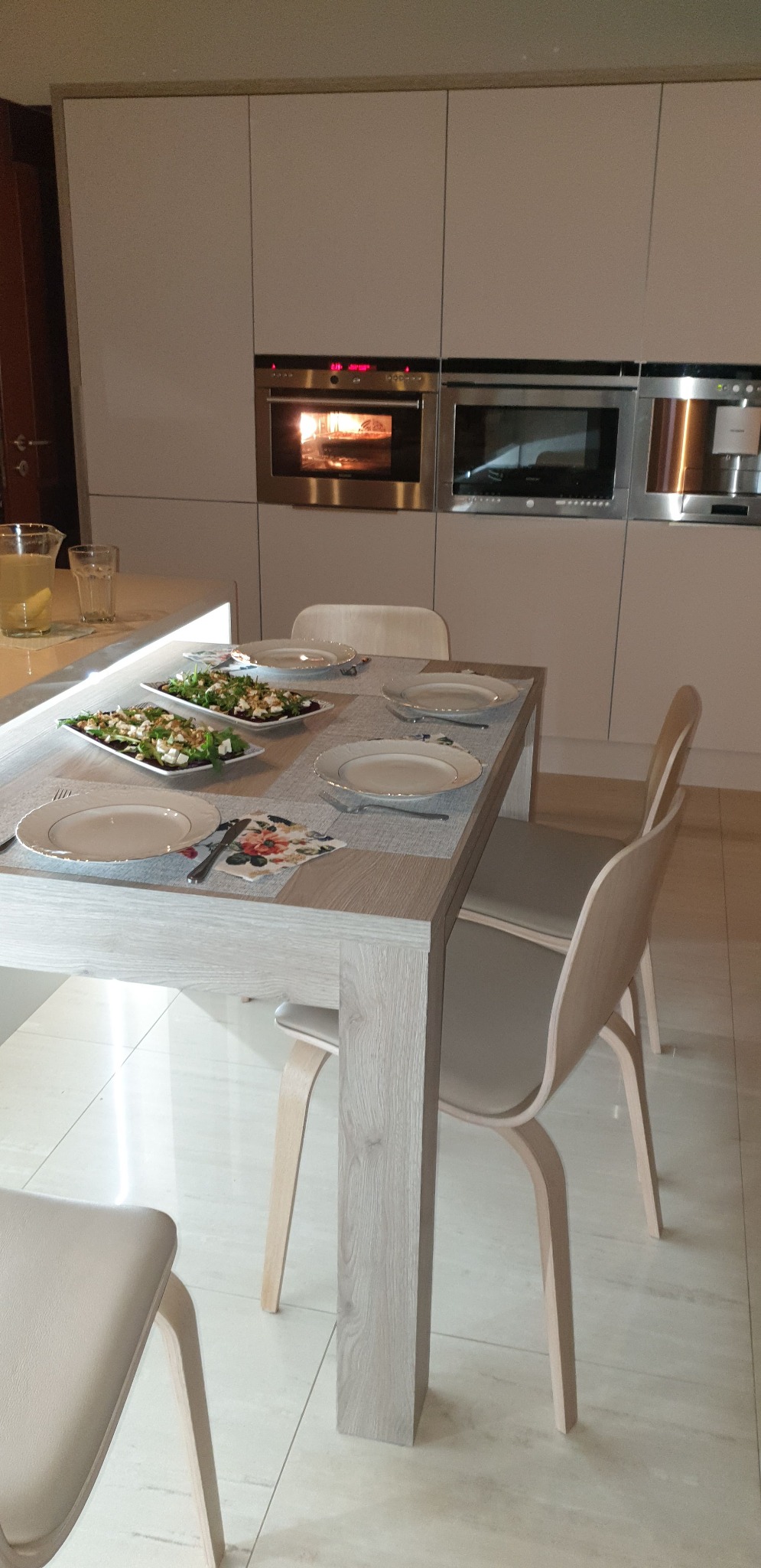 Nowoczesna kuchnia z zabudową w jasnych odcieniach, stół z nakryciem i sałatkami, piekarnik w zabudowie, minimalistyczny design.