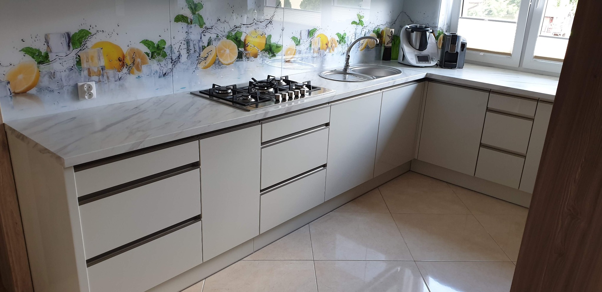 Jasna kuchnia z białymi frontami szafek, marmurowym blatem i szklanym panelem z motywem cytryn i lodu, widoczna płyta gazowa i zlewozmywak.