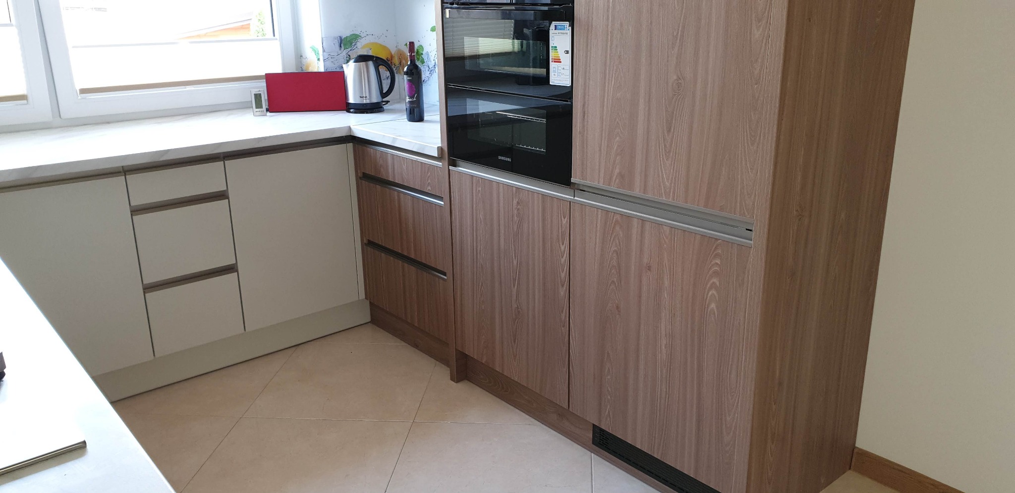 Nowoczesna zabudowa kuchenna z frontami w odcieniach beżu i brązu, z wbudowanym piekarnikiem i płytą grzewczą, uzupełniona o biały blat i akcesoria kuchenne.