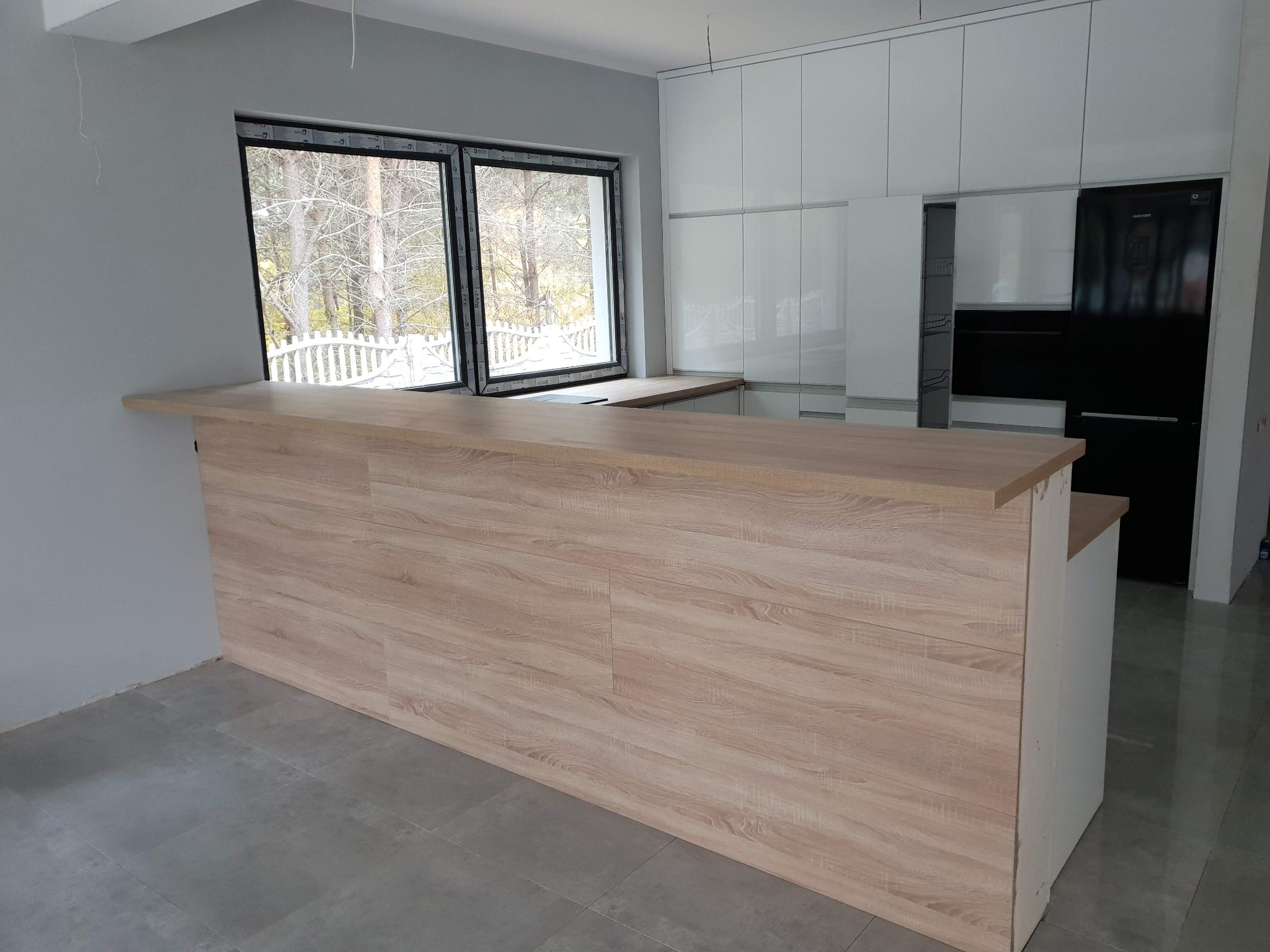 Jasna kuchnia z drewnianym blatem wyspowym i białymi szafkami, widok na zimowy krajobraz przez okno.