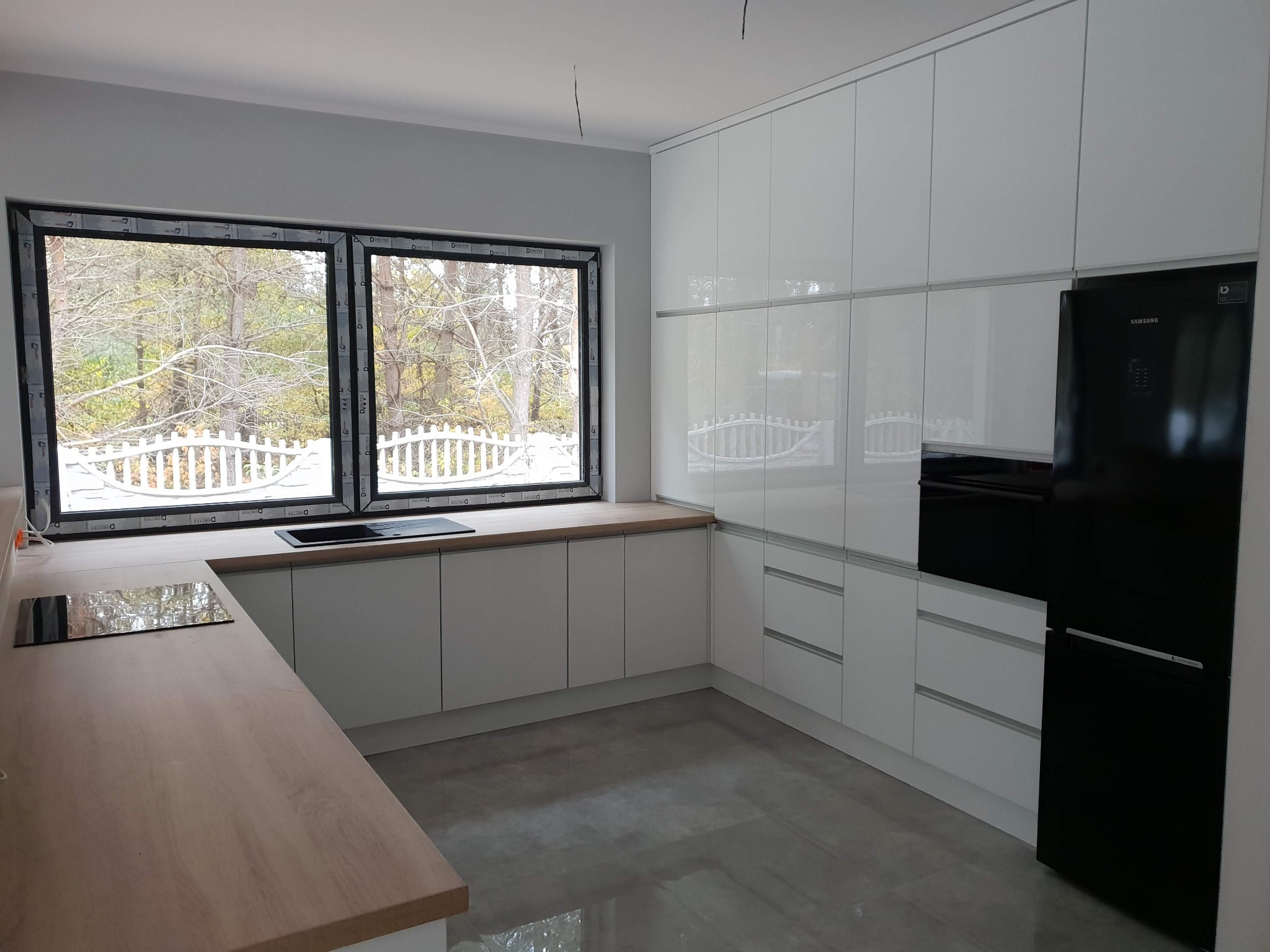 Jasna kuchnia z białymi, błyszczącymi frontami, drewnianym blatem i dużym oknem z widokiem na ogród, czarna lodówka Samsung po prawej stronie.