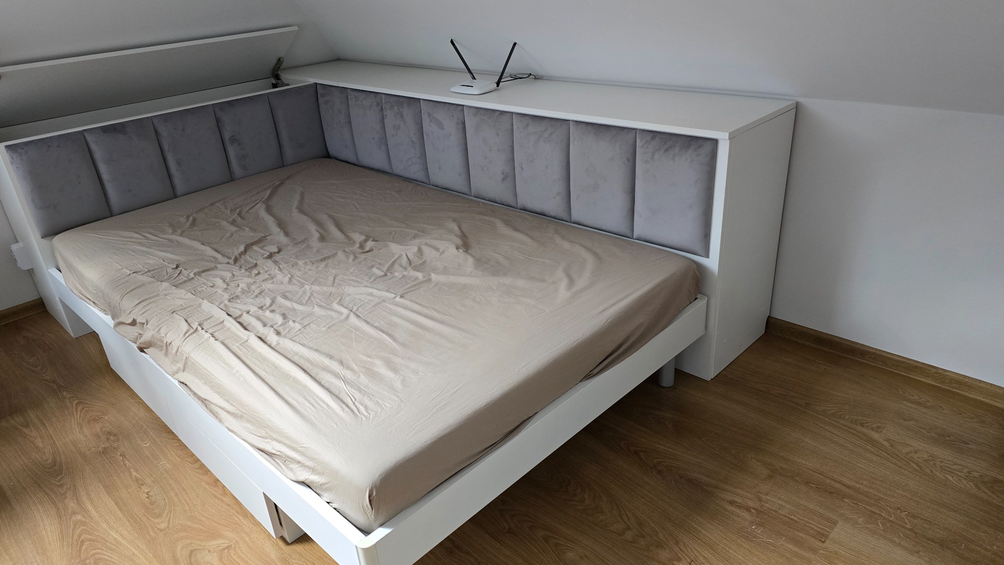 Białe łóżko na wymiar z szarym, tapicerowanym wezgłowiem w pionowe panele, z półką na górze, na której stoi router WiFi. Łóżko znajduje się na poddaszu.