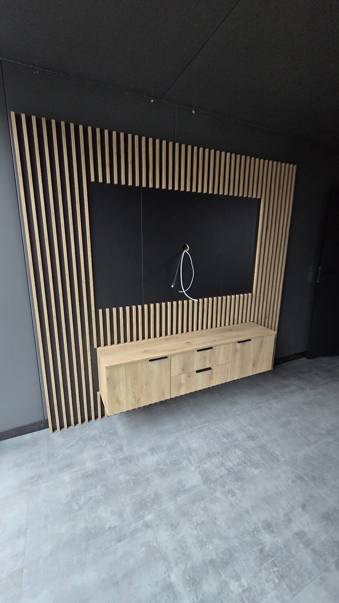 Nowoczesna zabudowa ściany z lamelami i szafką RTV z jasnego drewna, czarne tło pod telewizor z wystającym kablem, szara podłoga.