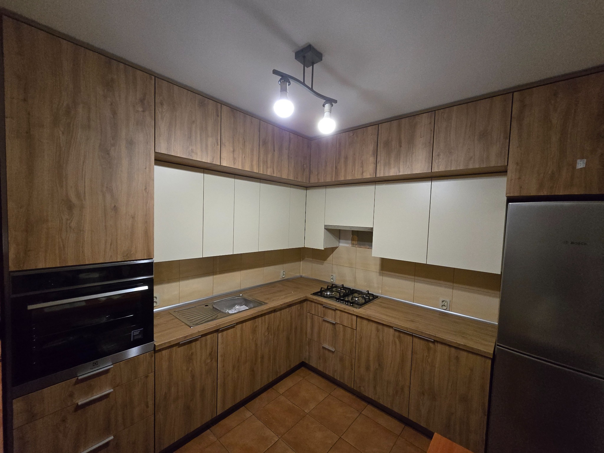 Nowoczesna kuchnia z drewnianymi szafkami dolnymi i białymi górnymi, oświetlona lampą sufitową z dwoma żarówkami, widoczny piekarnik Electrolux i lodówka Bosch.