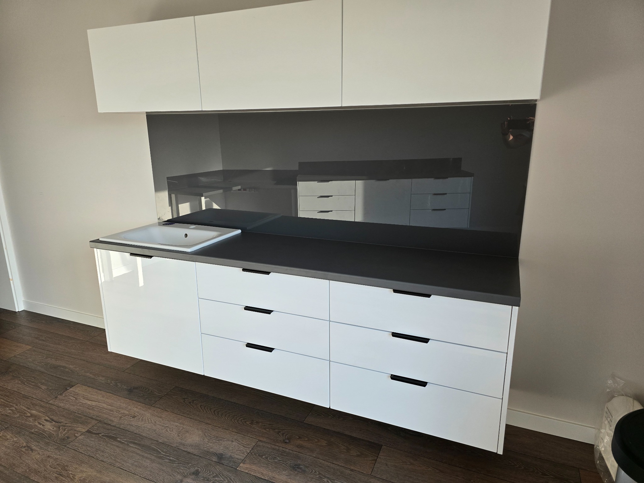 Nowoczesna zabudowa kuchenna z białymi frontami, czarnym blatem i lustrzanym panelem ściennym, w którym odbija się fragment innego pomieszczenia.