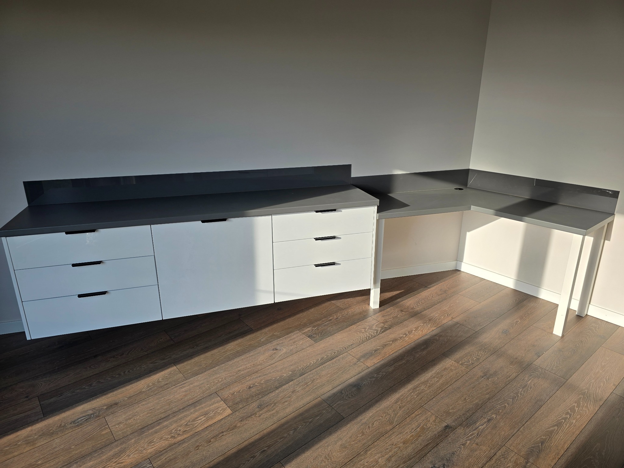 Białe szafki z szarym blatem i czarnymi uchwytami, tworzące narożne biurko na ciemnej drewnianej podłodze w pustym pomieszczeniu.