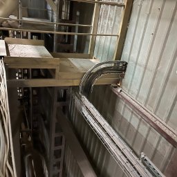 WAWEKO SPÓŁKA Z OGRANICZONĄ ODPOWIEDZIALNOŚCIĄ - Metalowa trasa kablowa, wypełniona przewodami, biegnąca wzdłuż ściany wewnątrz budynku, widoczna pod kątem z dołu.