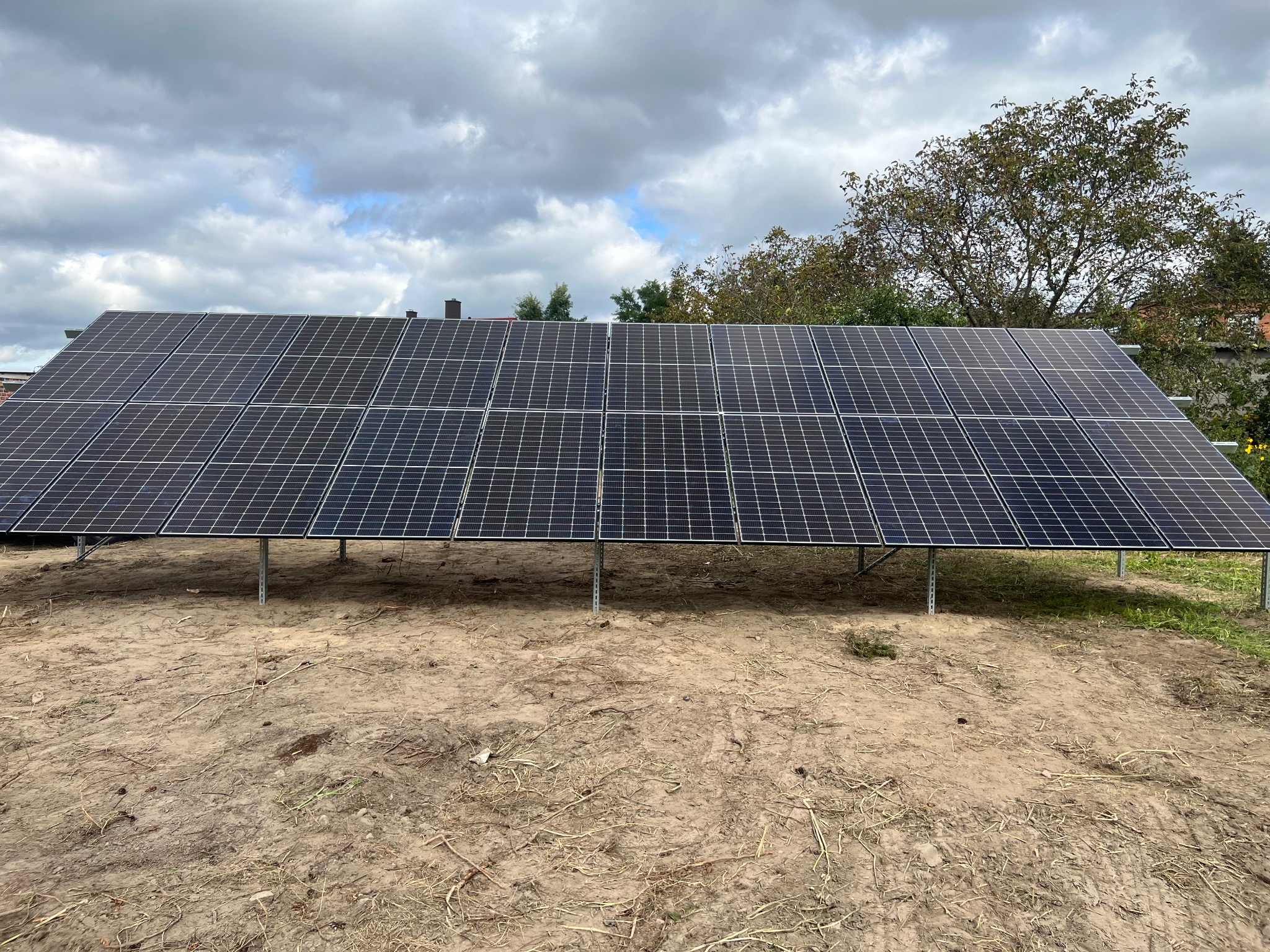 Panel słoneczny na gruncie, zamontowany na metalowej konstrukcji, z widocznymi chmurami na niebie i fragmentami drzew w tle. Teren wokół paneli ziemisty z resztkami roślinności.