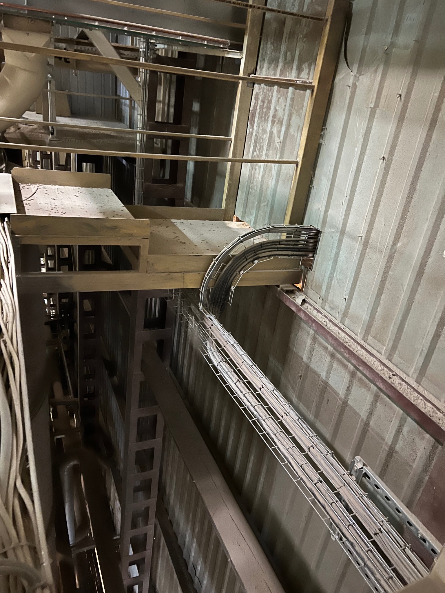 Metalowa trasa kablowa, wypełniona przewodami, biegnąca wzdłuż ściany wewnątrz budynku, widoczna pod kątem z dołu.