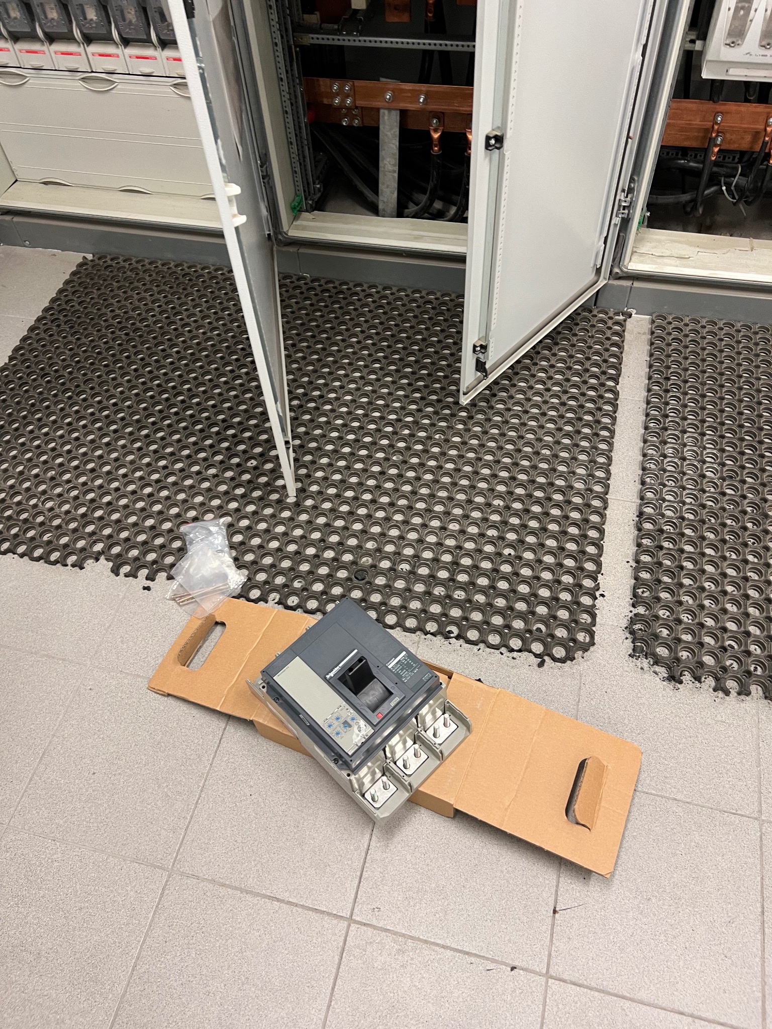 Wyjęty z opakowania wyłącznik elektryczny Schneider Electric na kartonowym podkładzie, leżący na podłodze z płytek w Żerkowie, obok otwartej szafy rozdzielczej z gumową wycieraczką. Widoczny...