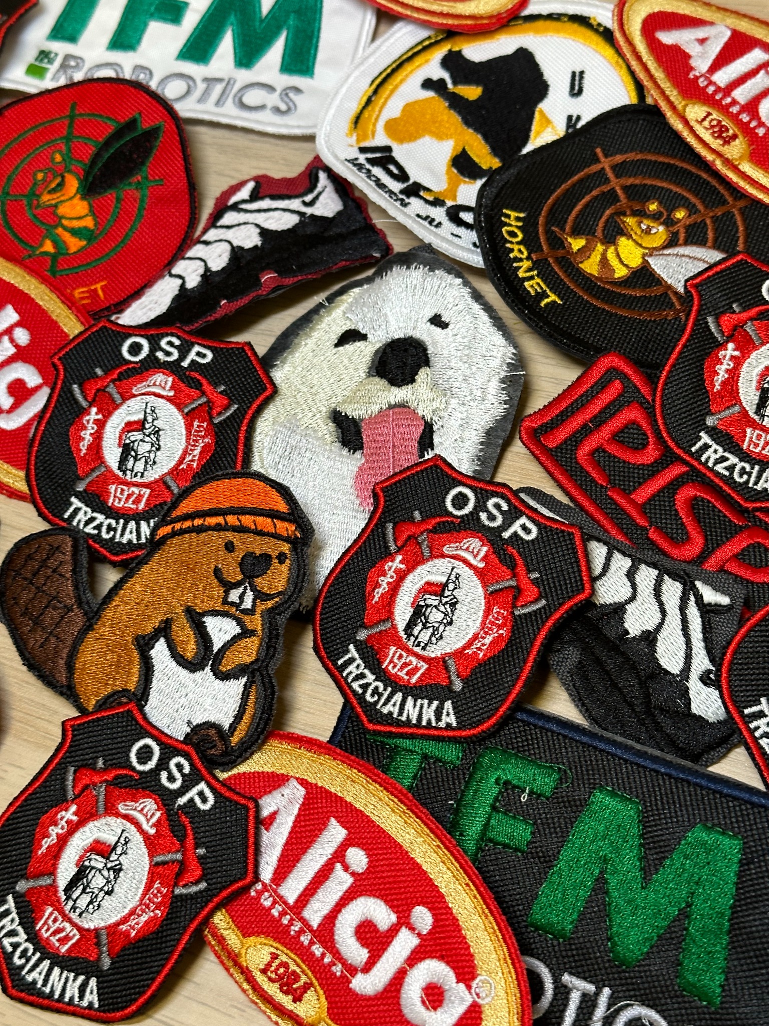 Kolekcja różnorodnych naszywek haftowanych, w tym motywy strażackie OSP Trzcianka, pszczoły Hornet, bobra w czapce i psa z wywieszonym językiem, ułożone na drewnianym podłożu.