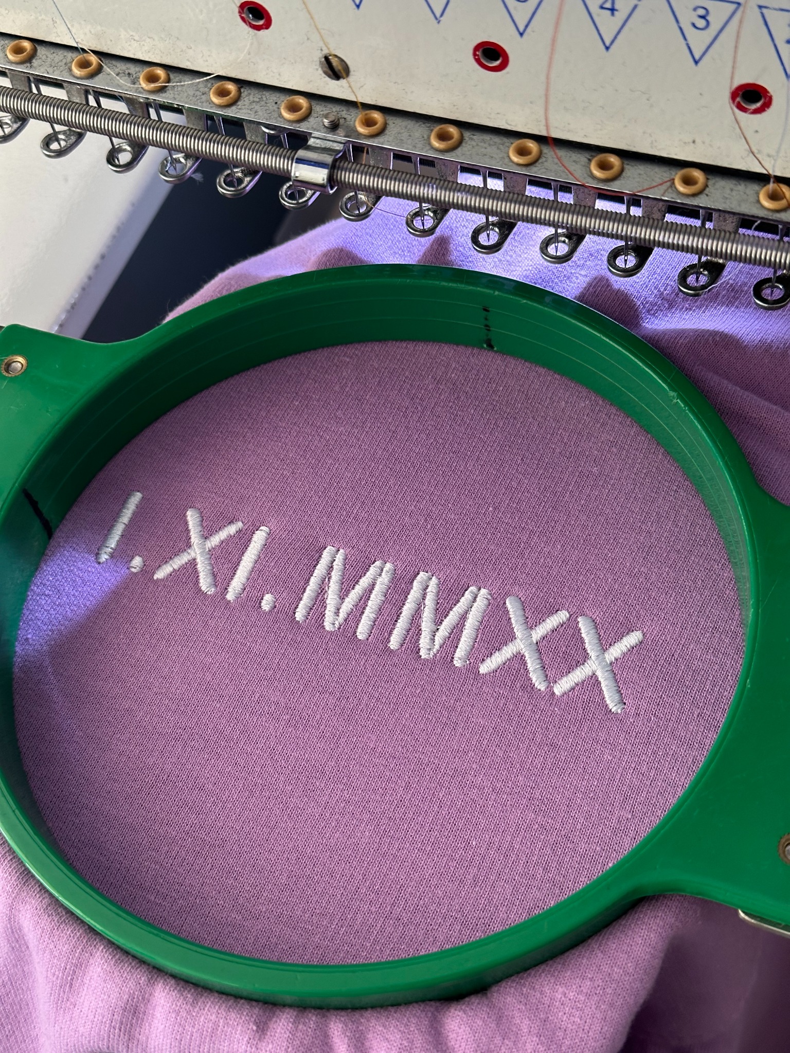 Proces haftowania daty 'I.XI.MMXX' białą nicią na fioletowej tkaninie, widoczne ramię maszyny hafciarskiej i zielona ramka stabilizująca materiał.