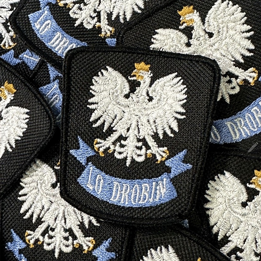 Zbliżenie na czarne, kwadratowe naszywki z wyhaftowanym białym orłem w koronie i niebieską wstęgą z napisem 'LO DROBIN', ułożone warstwami.
