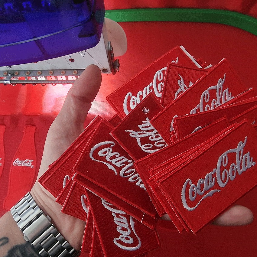 Dłoń trzymająca stos czerwonych naszywek z wyhaftowanym logo Coca-Cola, w tle maszyna do haftowania i wzory butelek Coca-Cola.