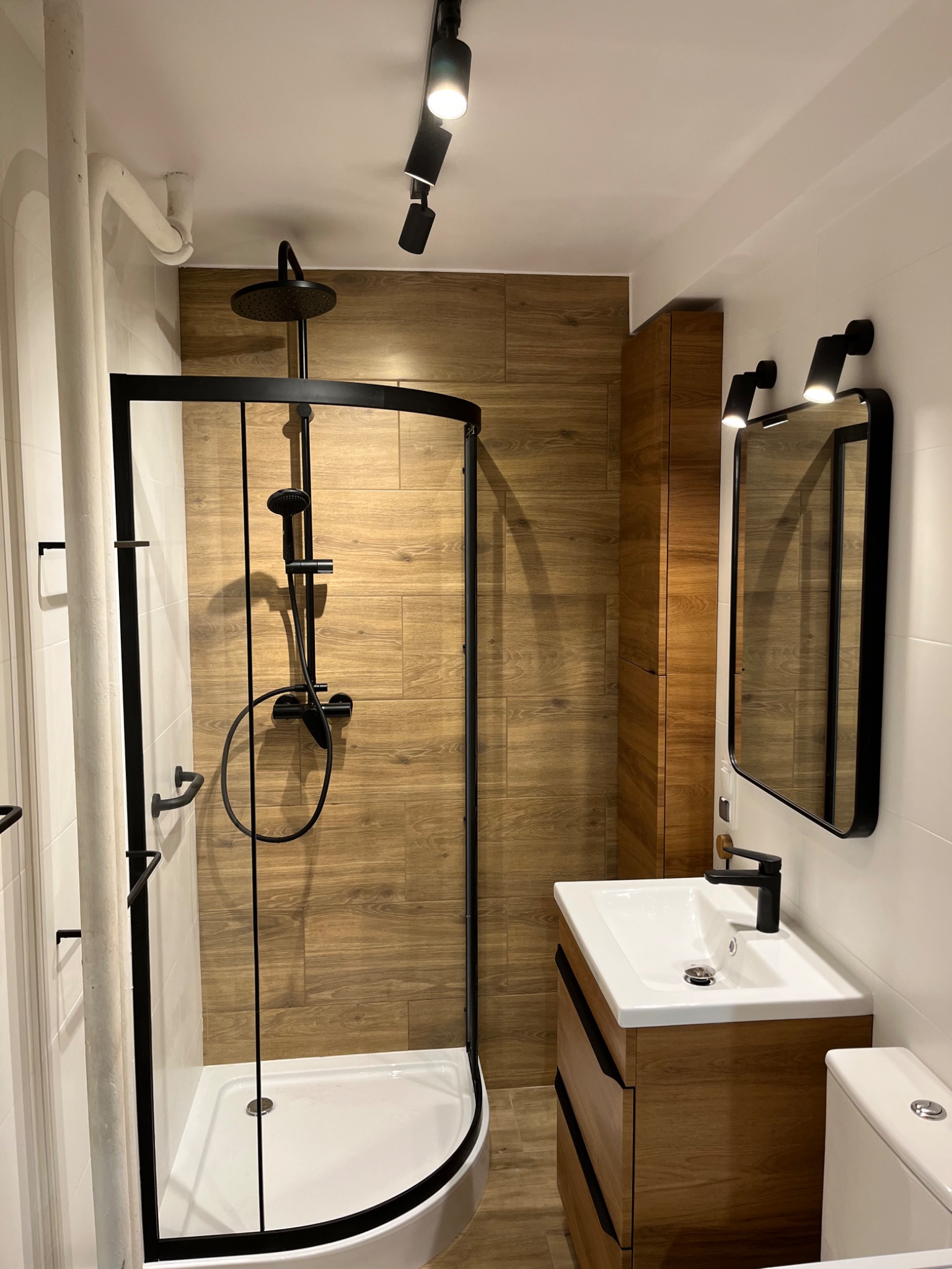 Nowoczesna łazienka z kabiną prysznicową z czarną ramą, drewnopodobnymi płytkami na ścianie prysznica, szafką z umywalką w odcieniach drewna z czarną armaturą i oświetleniem punktowym