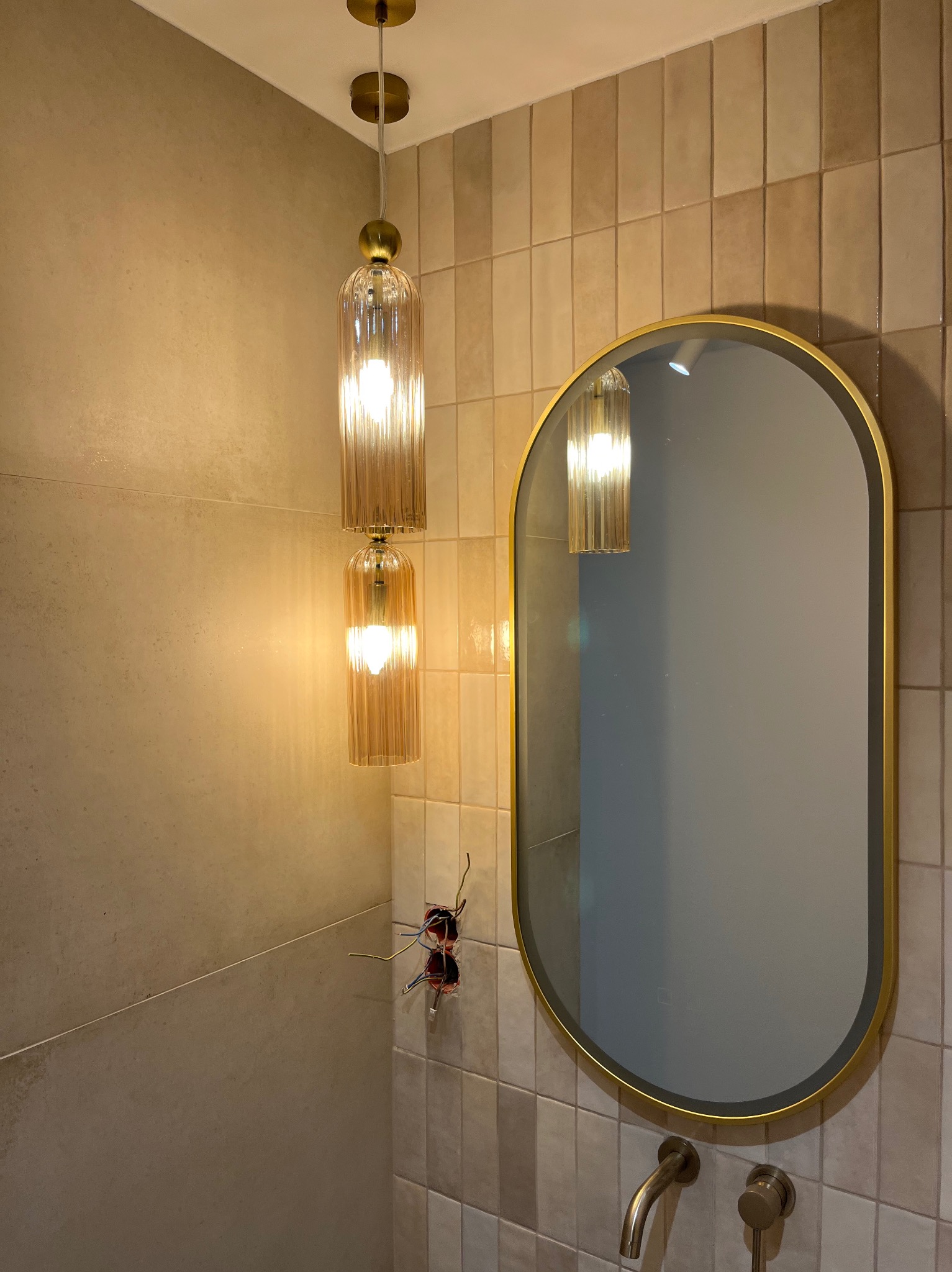 Nowoczesna łazienka w trakcie wykańczania: wisząca lampa z żłobionego szkła, owalne lustro w złotej ramie, bateria umywalkowa w kolorze szczotkowanego złota i odkryta instalacja elektryczna...
