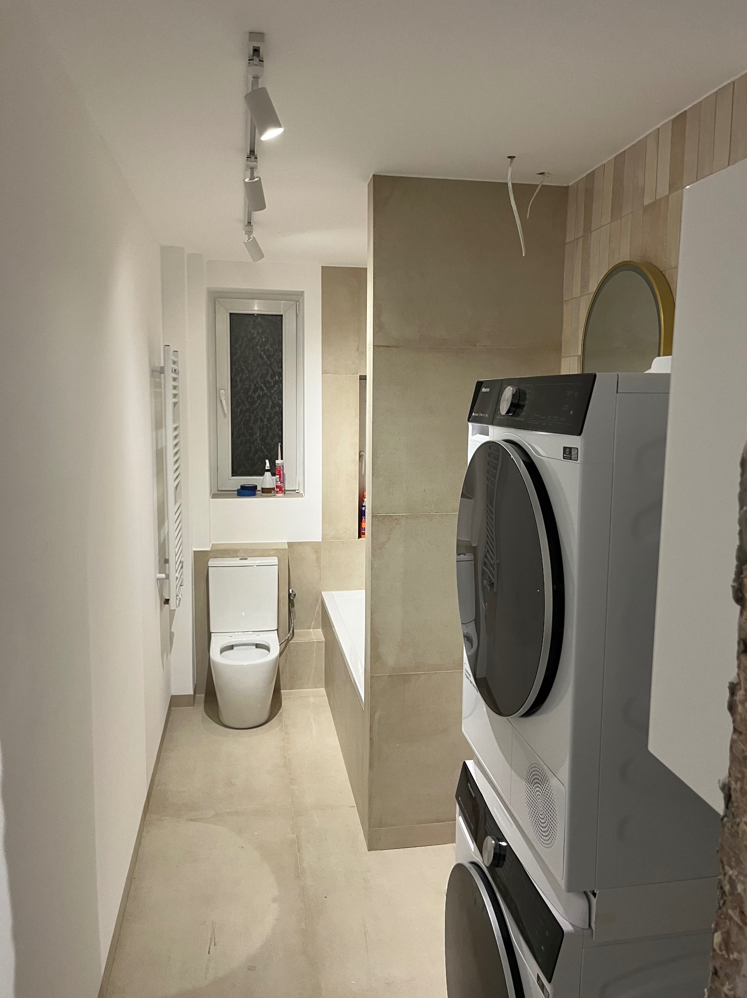 Nowoczesna łazienka w beżowych odcieniach z toaletą, grzejnikiem drabinkowym, wanną i pralko-suszarką Hisense w trakcie wykańczania, widoczne wystające kable z sufitu.