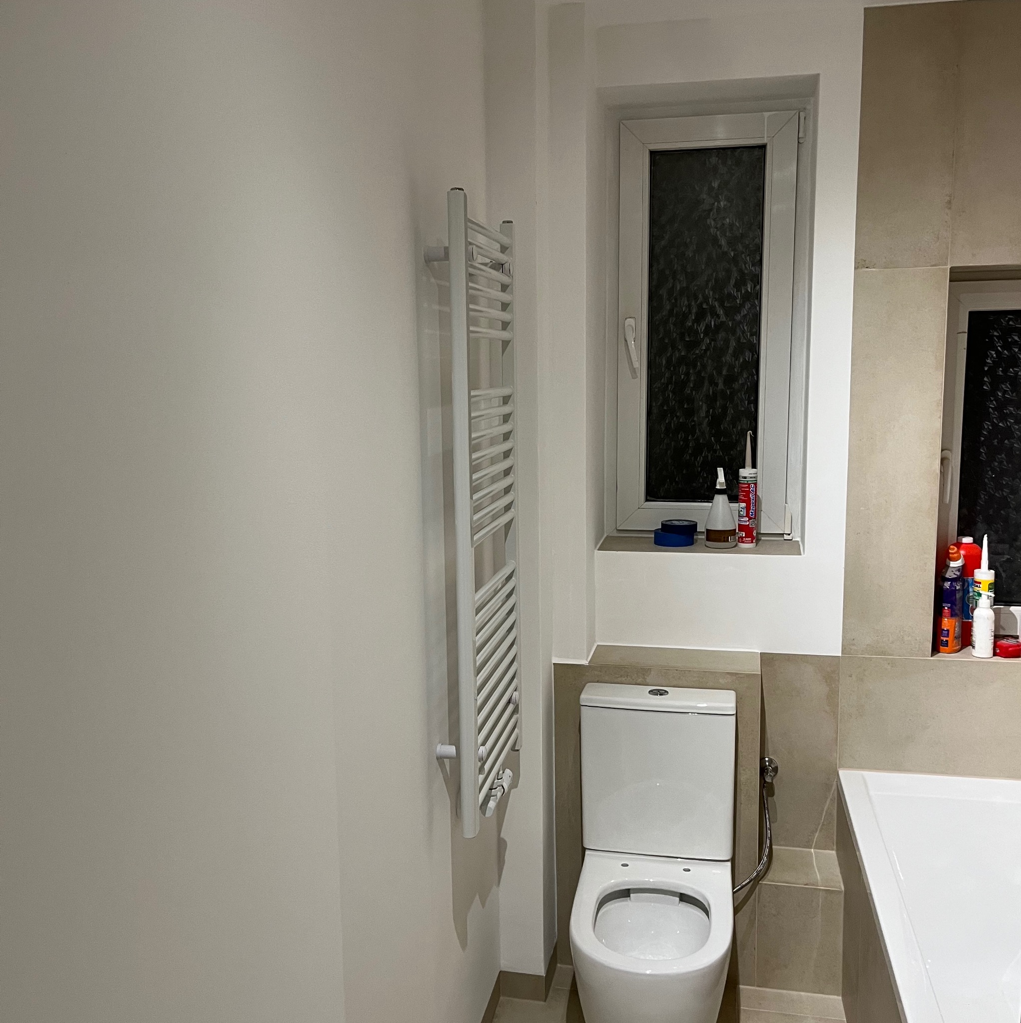 Nowoczesna, jasna łazienka po remoncie z białym grzejnikiem drabinkowym, toaletą i fragmentem wanny, uzupełniona o okno z akcesoriami montażowymi na parapecie.