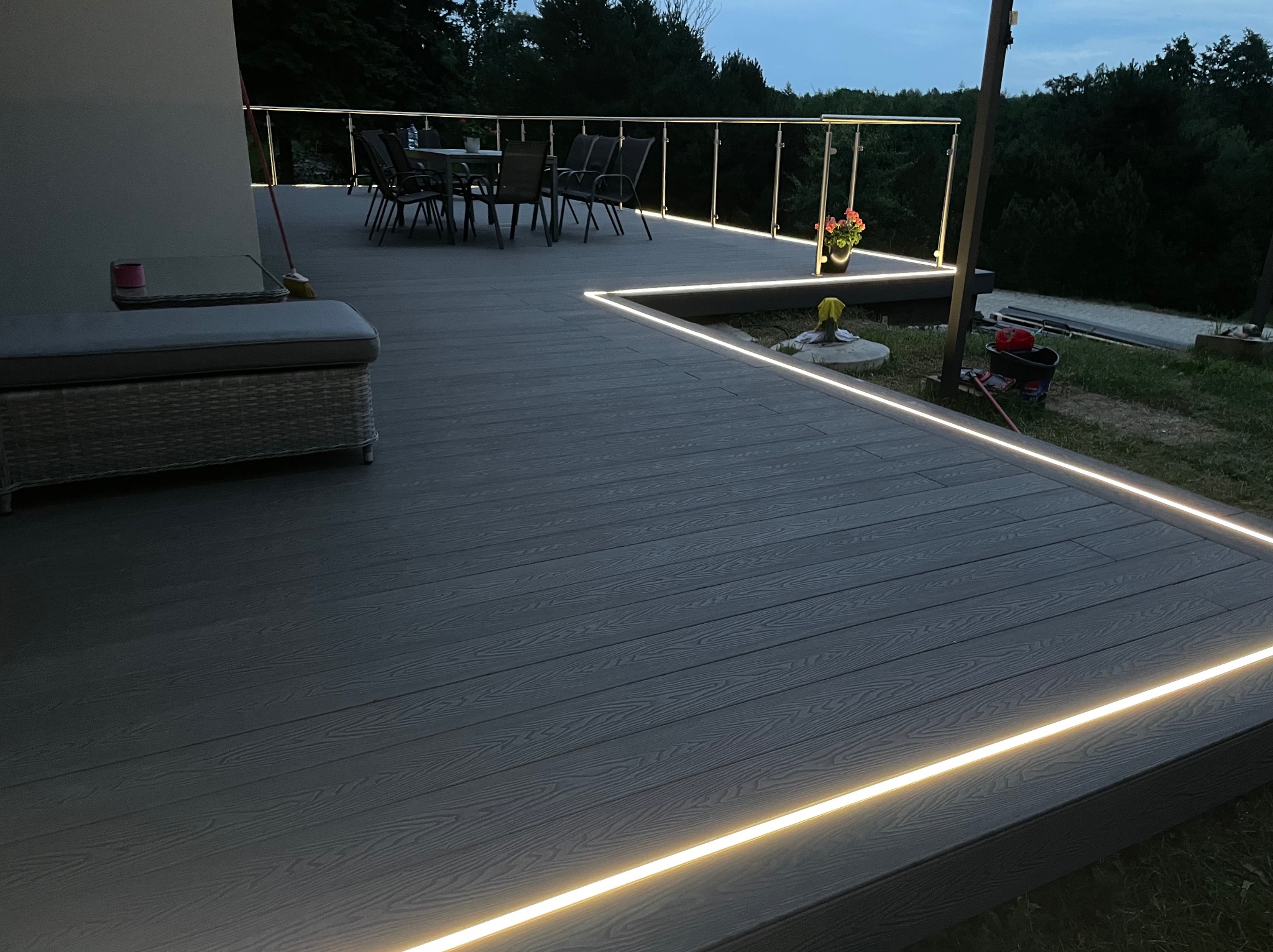 Nowoczesny taras z deski kompozytowej oświetlony liniowym światłem LED, z balustradą ze stali nierdzewnej i szklanym wypełnieniem. Na tarasie stół z krzesłami i doniczka z kwiatami. Widoczne...