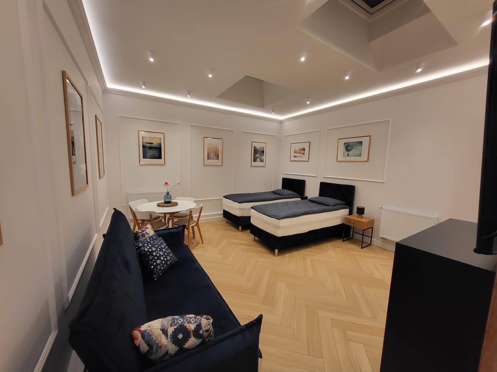 Jasne wnętrze pokoju z parkietem w jodełkę, dwoma pojedynczymi łóżkami, sofą z poduszkami, okrągłym stołem z krzesłami i dekoracyjnymi obrazami na ścianach z listwami.