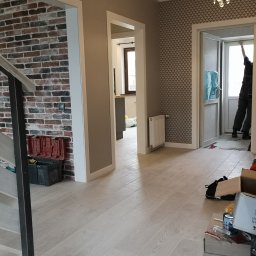 VOLHA PAVIADAIKA - Jasne panele podłogowe w korytarzu z widocznymi narzędziami i materiałami budowlanymi, ceglana ściana dekoracyjna po lewej, tapeta w geometryczny wzór po prawej stronie, otwarte drzwi w tle...