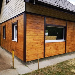 VOLHA PAVIADAIKA - Drewniana elewacja domu z widocznymi oknami i brązowymi ramami wykończeniowymi, jasne panele na górze, brązowy dach, częściowo widoczna osoba pracująca na zewnątrz.