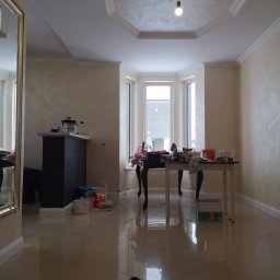 VOLHA PAVIADAIKA - Pokój po nałożeniu gładzi dekoracyjnej na ścianach i suficie, widoczny stół z narzędziami i pojemnikami, podłoga z połyskiem, centralnie umieszczona lampa wisząca z żarówką.