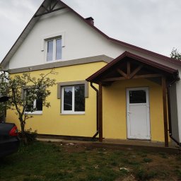 VOLHA PAVIADAIKA - Żółto-biały dom jednorodzinny z nową elewacją, widoczne okna PCV i drewniana weranda z brązowym dachem, obok kwitnące drzewo i czarny samochód z otwartym bagażnikiem.