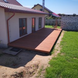 VOLHA PAVIADAIKA - Nowoczesny taras z desek kompozytowych przylegający do domu z czerwonym dachem, widoczny fragment ogrodu z wiszącym fotelem i budynkiem gospodarczym z pustaków.