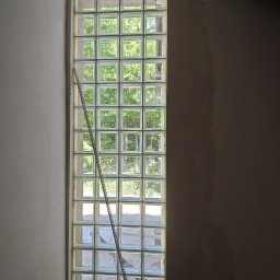 VOLHA PAVIADAIKA - Wysoki, wąski świetlik z luksferów w surowym wnętrzu, widoczna drabina oparta o szybę i stary grzejnik elektryczny u dołu