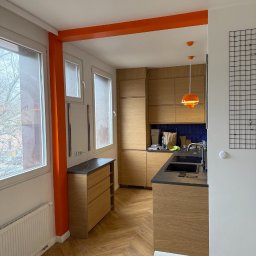 VOLHA PAVIADAIKA - Nowoczesna kuchnia z drewnianymi szafkami i blatem w kolorze grafitu, parkiet ułożony w jodełkę, pomarańczowe akcenty na ścianach i lampach, widok z perspektywy wejścia.