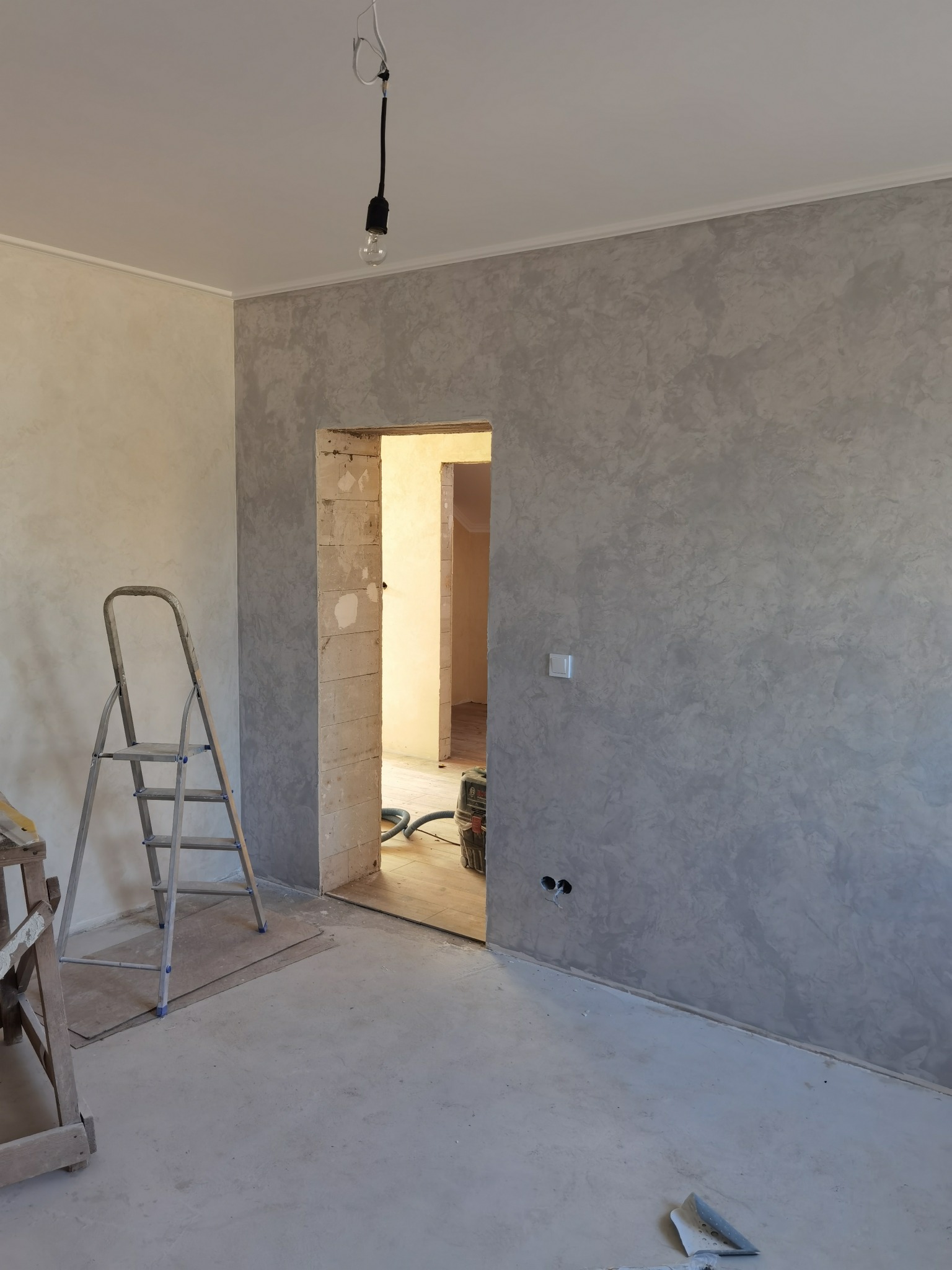 Pokój w trakcie prac wykończeniowych: ściana z szarą fakturą, surowy beton na podłodze, drabina aluminiowa, widoczne narzędzia i otwór drzwiowy prowadzący do kolejnego pomieszczenia z odkurzaczem.