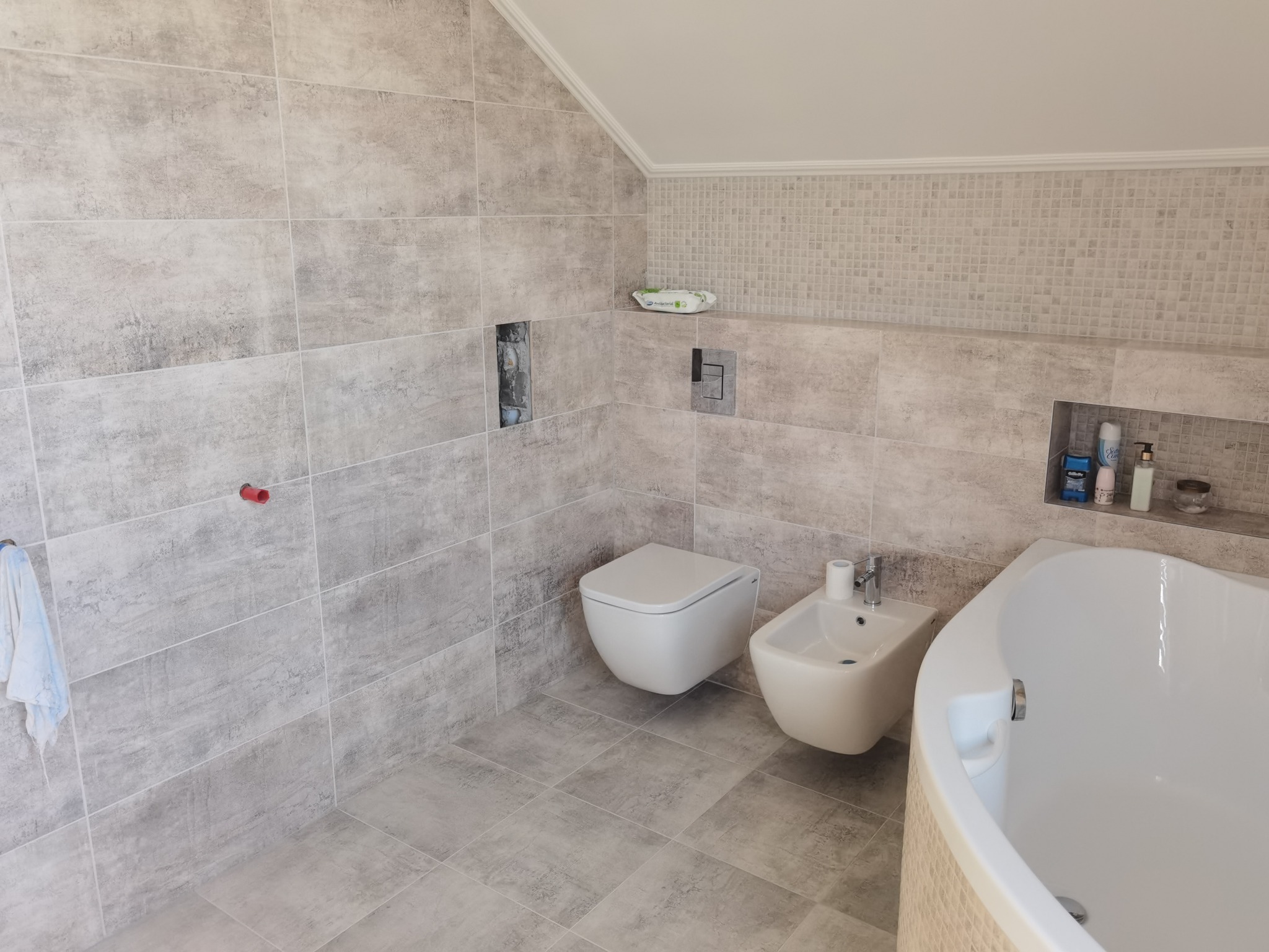 Nowoczesna łazienka w szarościach z wanną, bidetem i toaletą podwieszaną, wykończona płytkami imitującymi beton, z wnęką na kosmetyki i akcesoria.