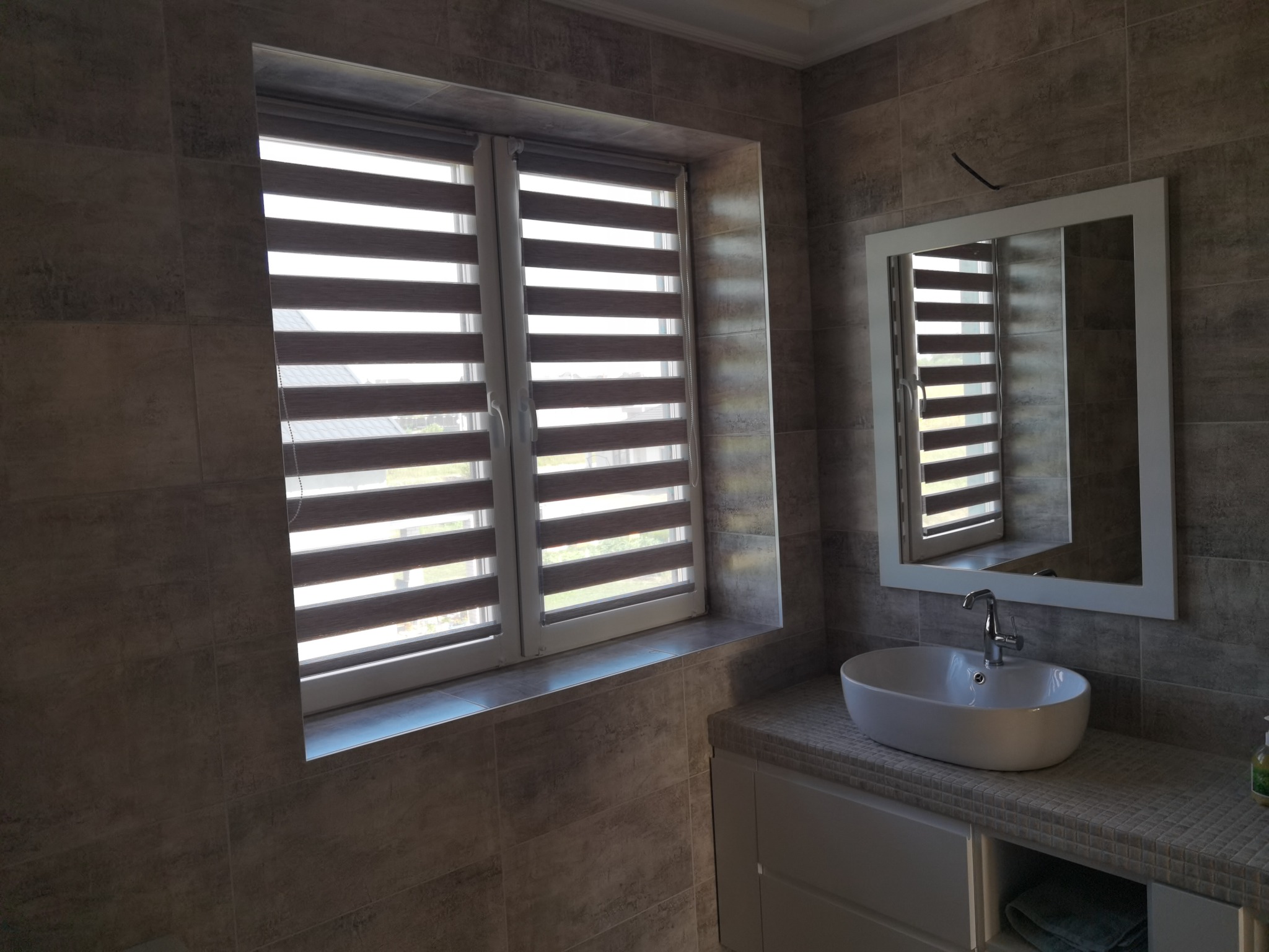 Nowoczesna łazienka z szarymi płytkami imitującymi kamień, białą umywalką nablatową, lustrem w białej ramie i oknem z roletami dzień-noc.
