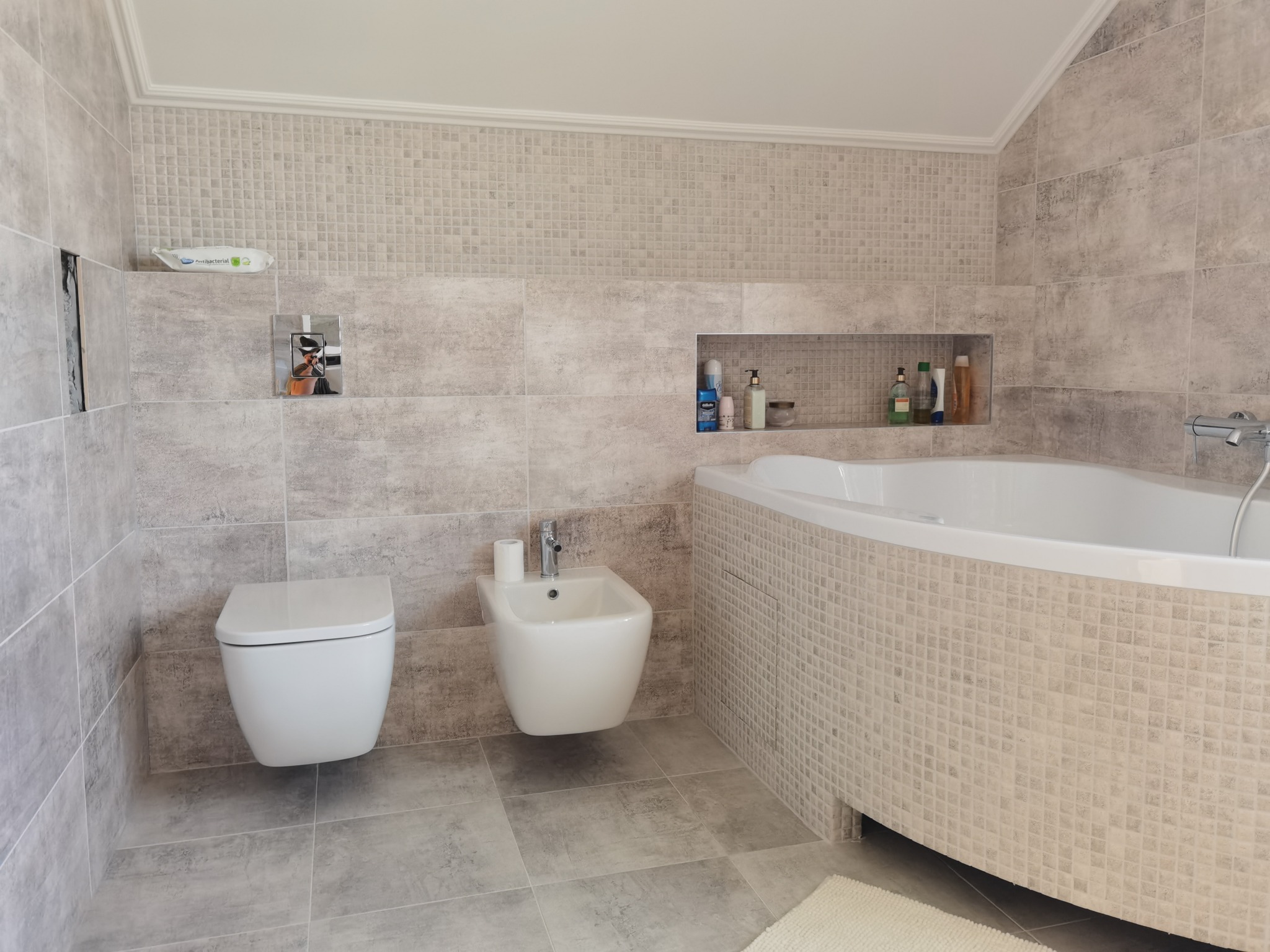 Nowoczesna łazienka w odcieniach szarości z wiszącą toaletą, bidetem, wanną obudowaną mozaiką i wnęką na kosmetyki.