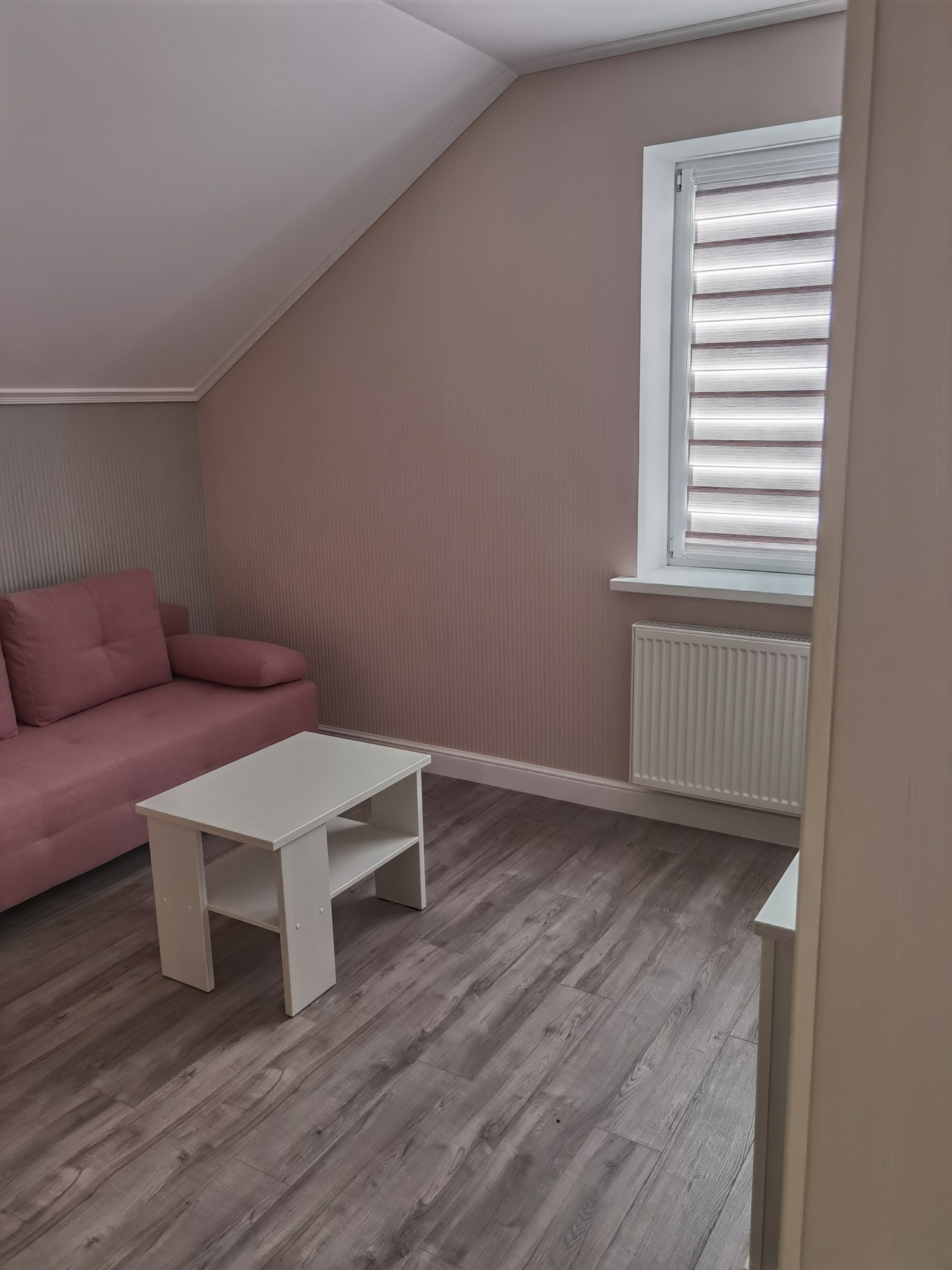 Pokój na poddaszu z drewnianą podłogą, różową sofą i białym stolikiem kawowym, ściany pomalowane na delikatny różowy kolor, okno z roletami dzień-noc.