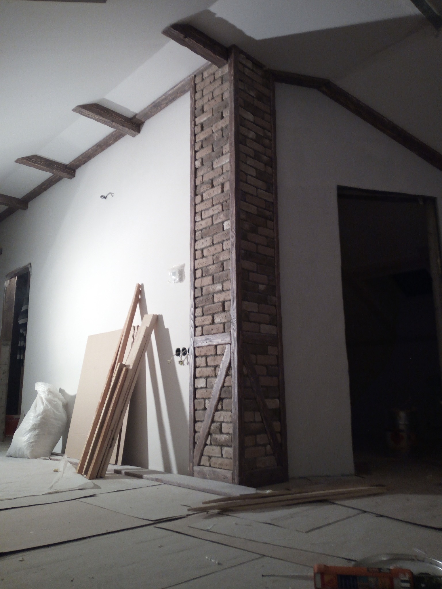 Wnętrze w trakcie prac wykończeniowych: kolumna z cegły klinkierowej w drewnianej ramie, drewniane belki stropowe, ściany przygotowane do malowania, płyty i listwy przypodłogowe opierające się...