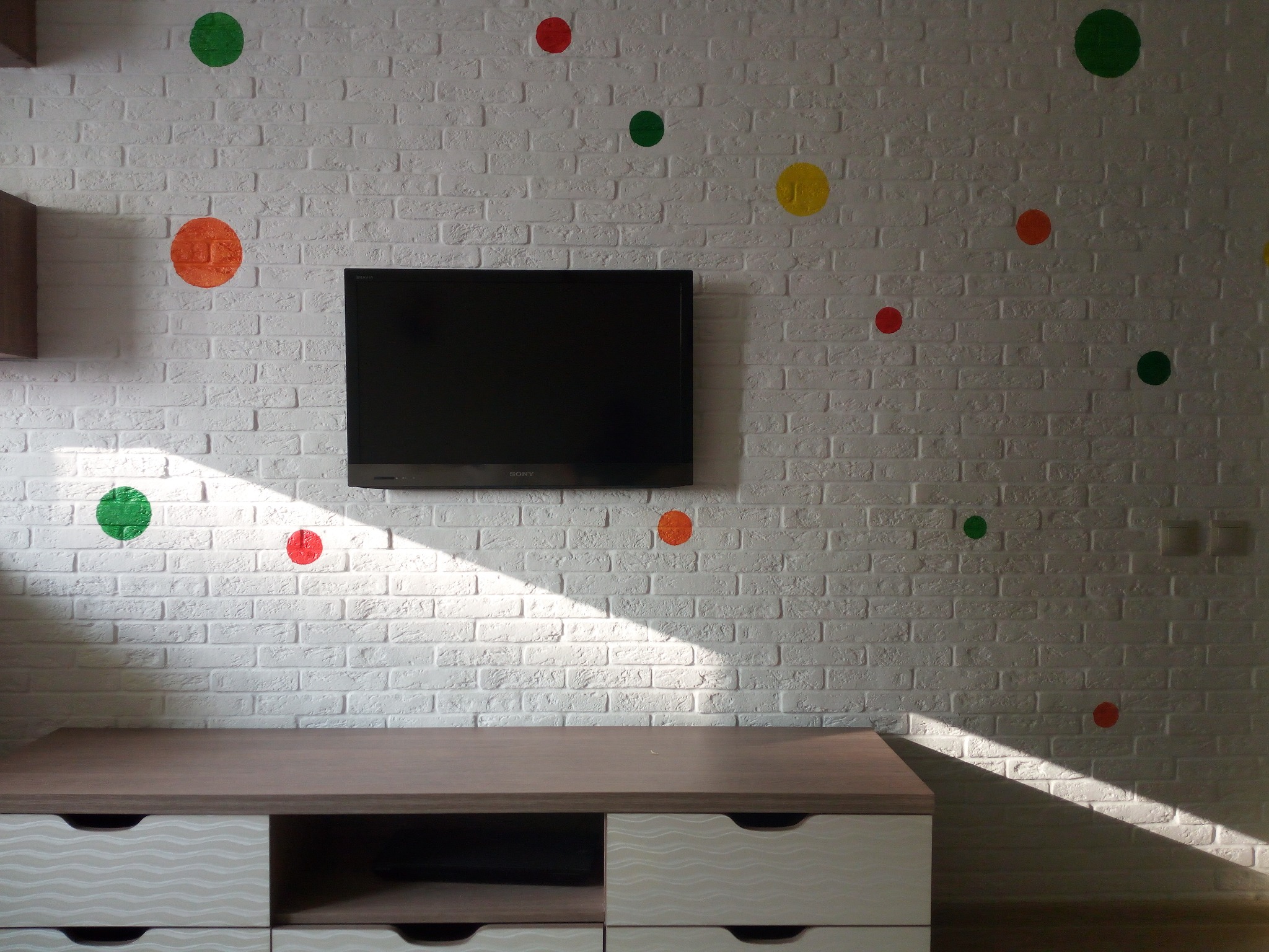 Biała ściana z cegły pomalowana na biało, udekorowana kolorowymi naklejkami w kształcie kropek, podwieszony telewizor i szafka RTV z szufladami w dolnej części, oświetlona popołudniowym słońcem.