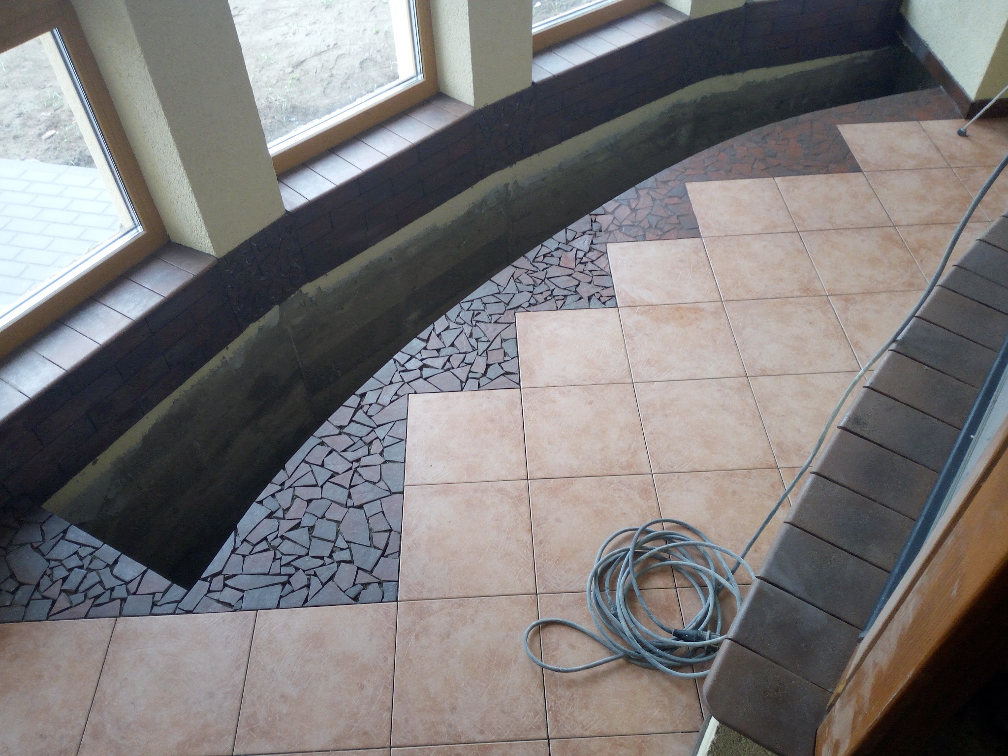 Układanie beżowych płytek podłogowych w połączeniu z mozaiką z drobnych, nieregularnych elementów w strefie okiennej; widoczny zwój kabla elektrycznego na podłodze.