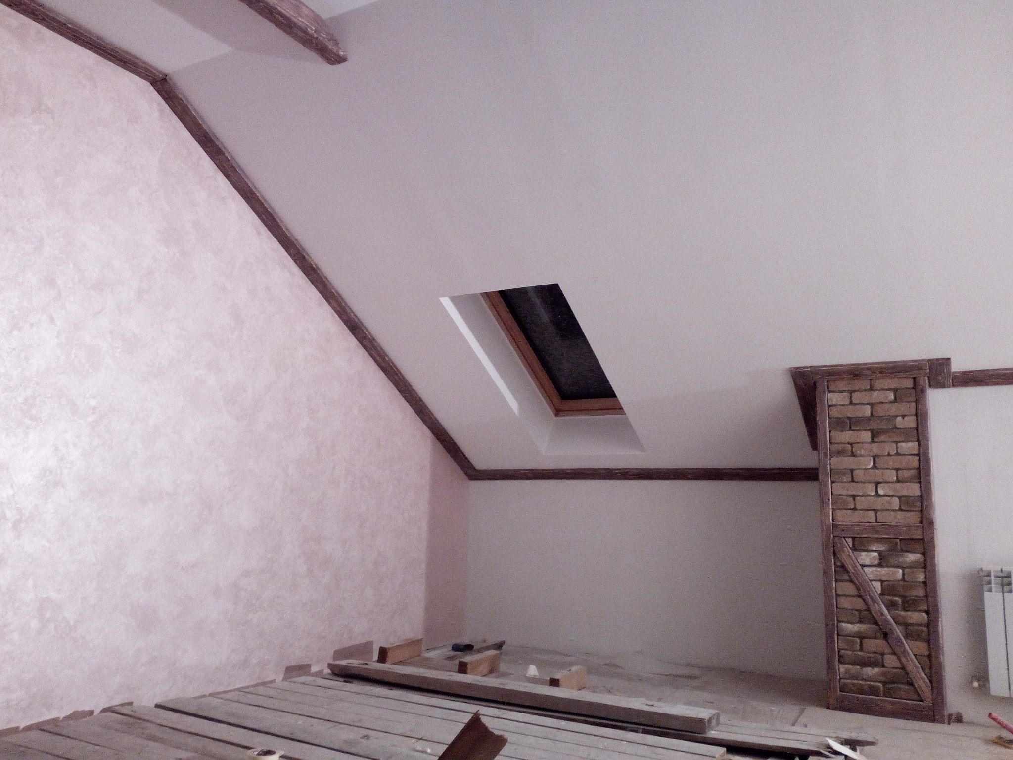 Poddasze w trakcie prac wykończeniowych, ściana pokryta dekoracyjną, różową fakturą, drewniane belki stropowe i obramowanie okna dachowego, surowa podłoga z desek, fragment ściany z cegły...