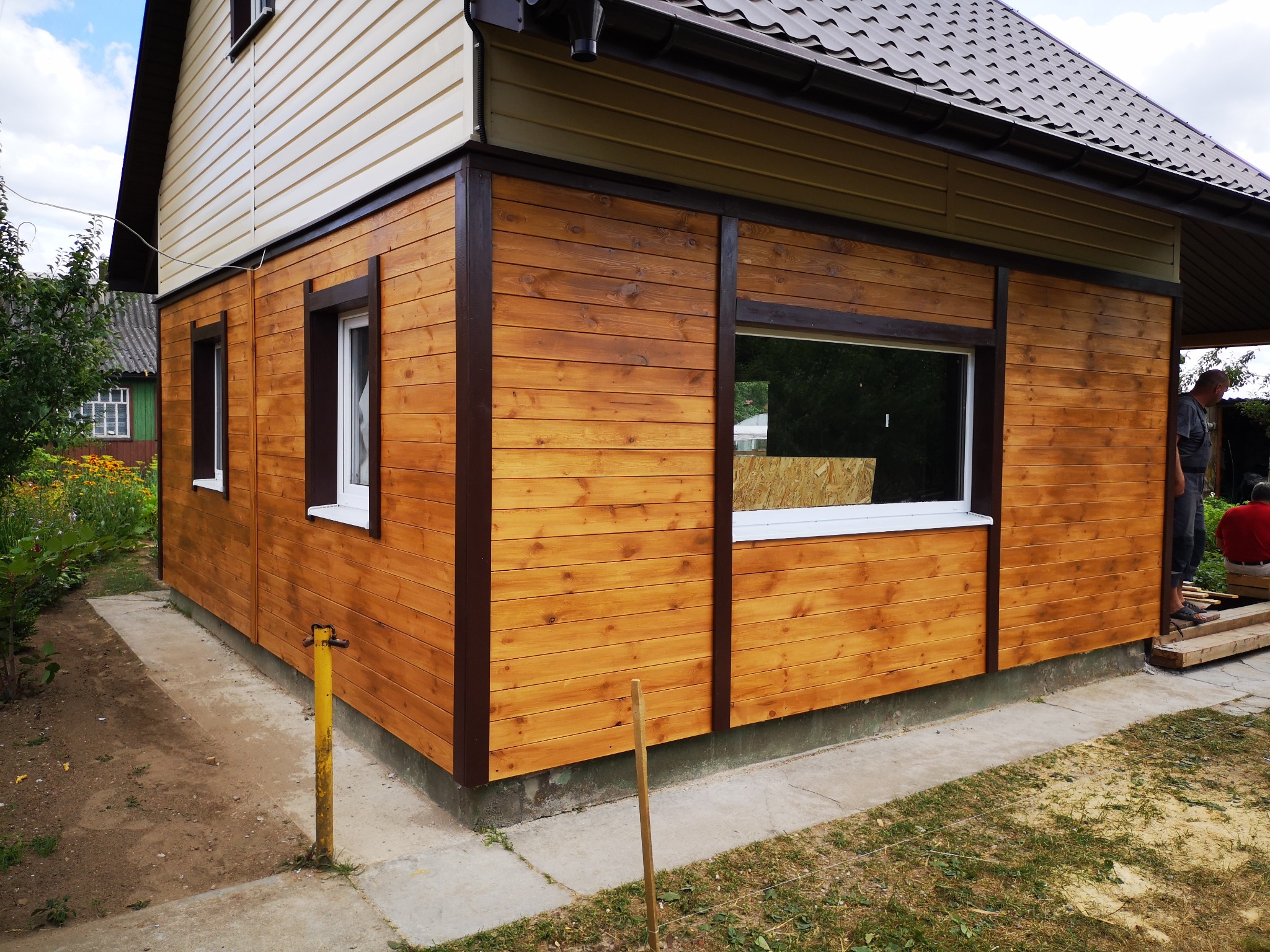 Drewniana elewacja domu z widocznymi oknami i brązowymi ramami wykończeniowymi, jasne panele na górze, brązowy dach, częściowo widoczna osoba pracująca na zewnątrz.