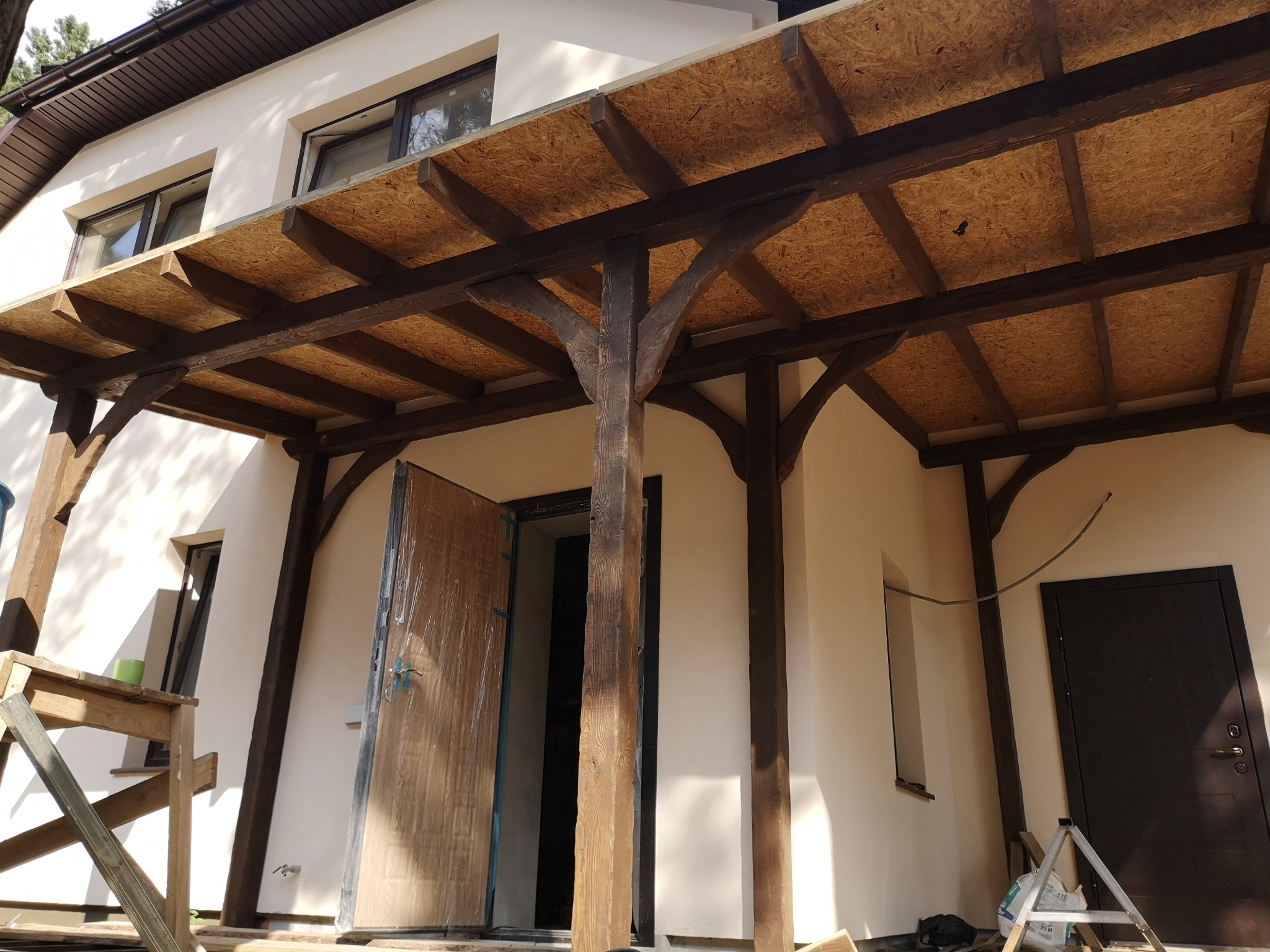 Zadaszenie tarasu z drewnianą konstrukcją i płytami OSB, widok na białą elewację budynku z oknami i drzwiami, częściowo zakryte folią ochronną, w tle drzewa.