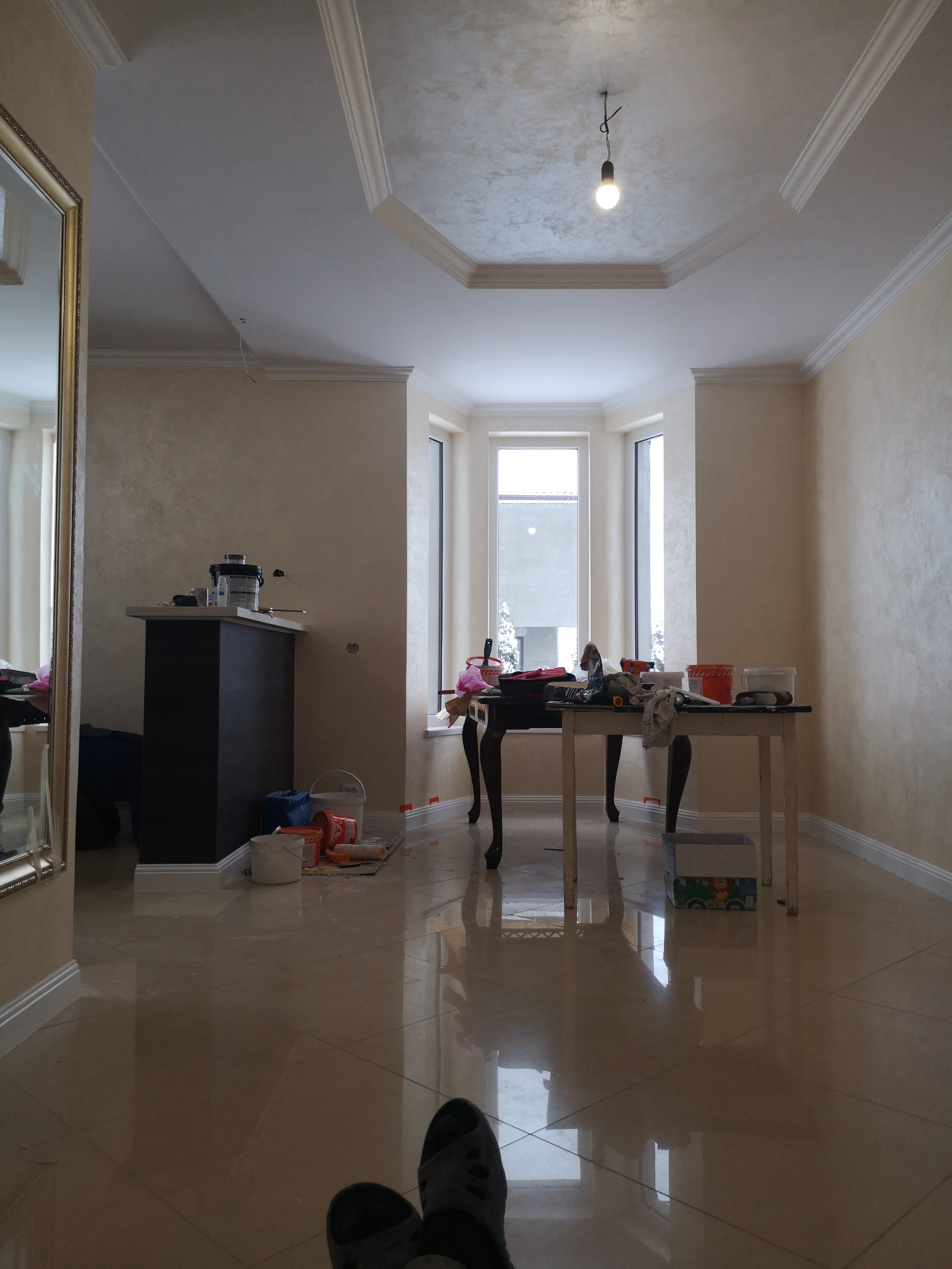 Pokój po nałożeniu gładzi dekoracyjnej na ścianach i suficie, widoczny stół z narzędziami i pojemnikami, podłoga z połyskiem, centralnie umieszczona lampa wisząca z żarówką.