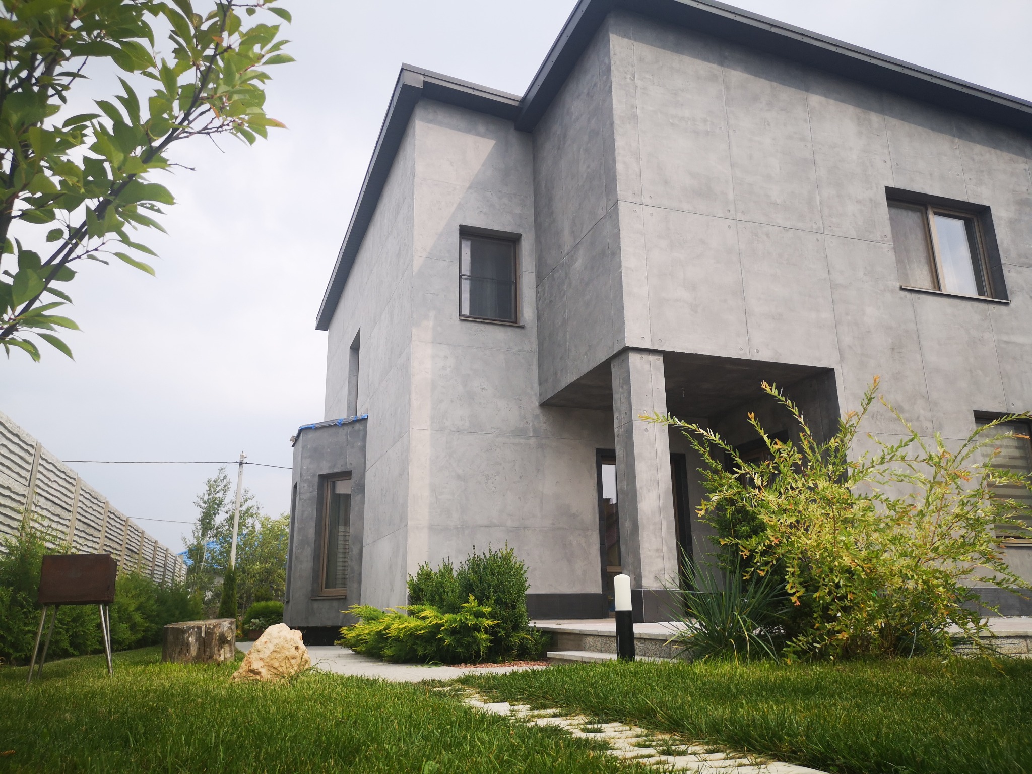 Nowoczesny dom jednorodzinny z szarą elewacją imitującą beton, widok z ogrodu z kamienną ścieżką i roślinnością.
