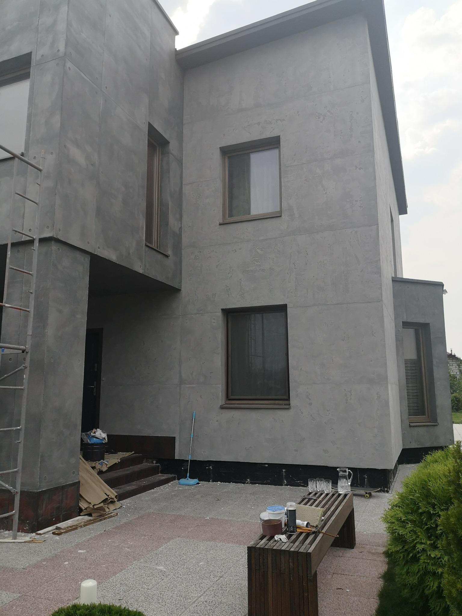 Elewacja domu z szarą, dekoracyjną gładzią, widoczne okna z brązowymi ramami, podest z narzędziami i akcesoriami malarskimi, częściowo widoczne rusztowanie.