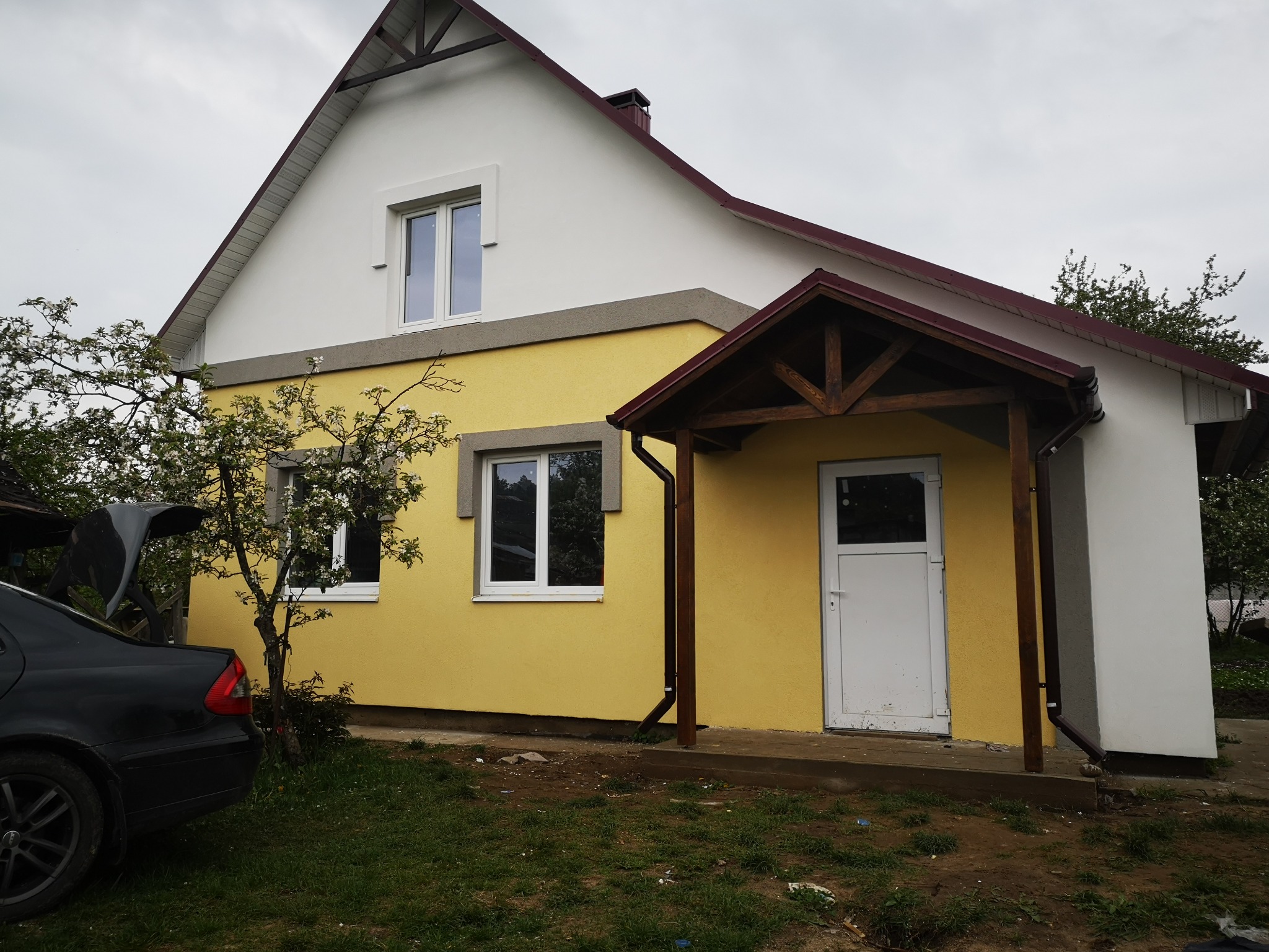 Żółto-biały dom jednorodzinny z nową elewacją, widoczne okna PCV i drewniana weranda z brązowym dachem, obok kwitnące drzewo i czarny samochód z otwartym bagażnikiem.