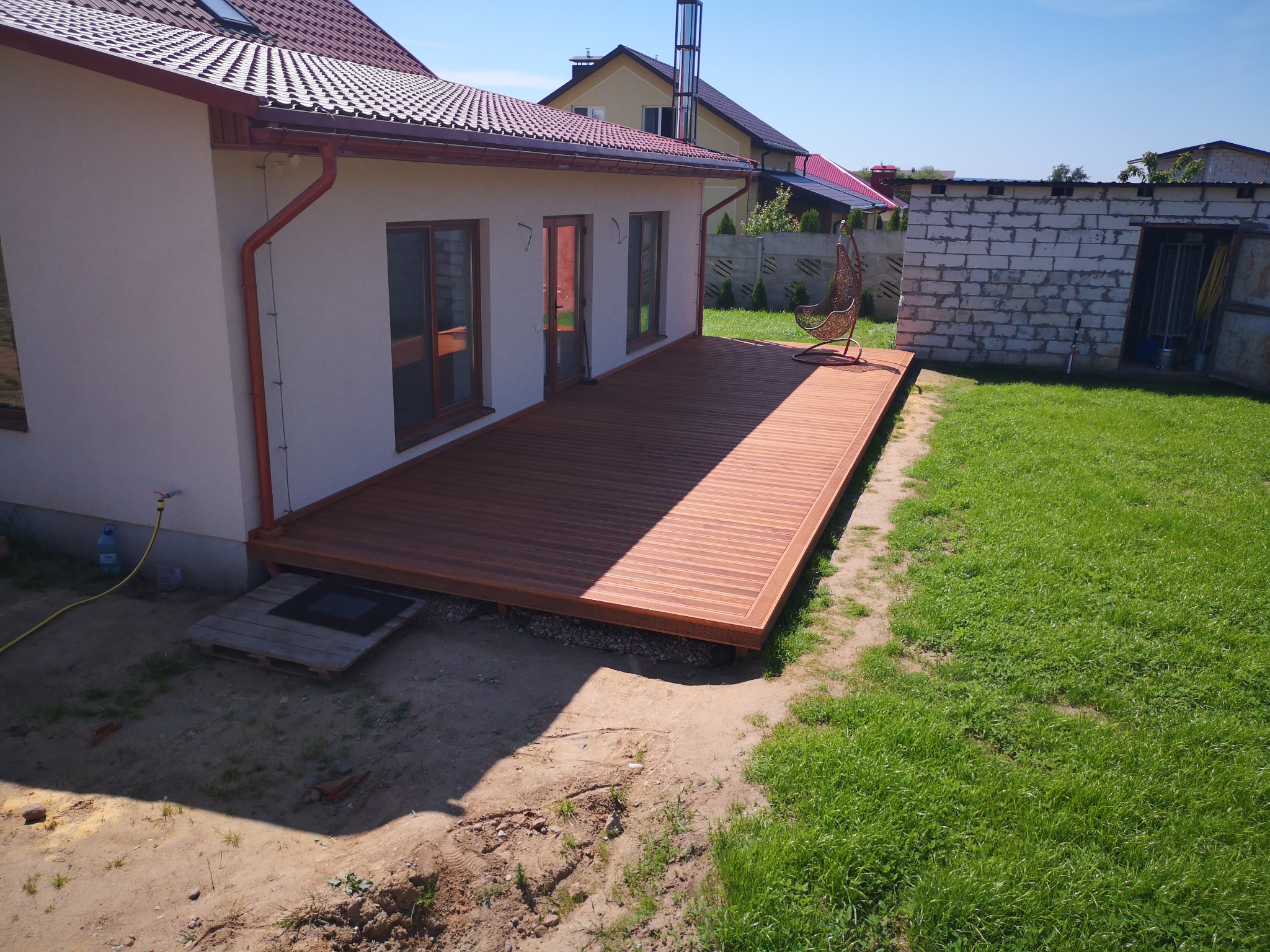 Nowoczesny taras z desek kompozytowych przylegający do domu z czerwonym dachem, widoczny fragment ogrodu z wiszącym fotelem i budynkiem gospodarczym z pustaków.